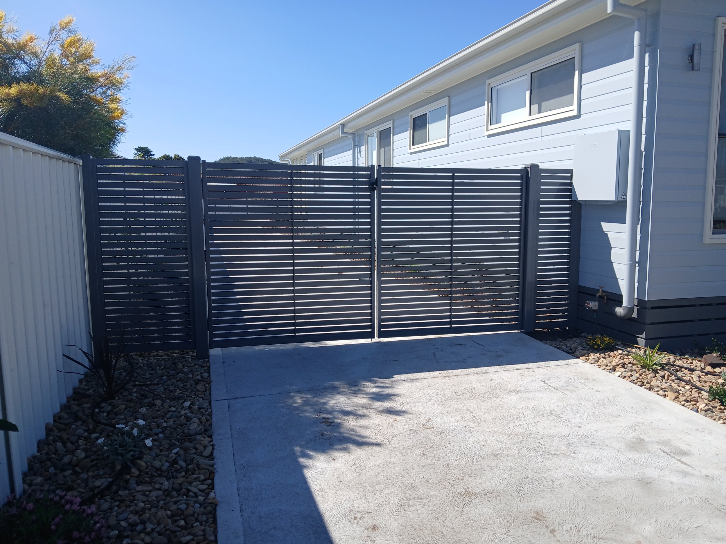 Black metal gate with horizontal slats in front of a white house with multiple windows, a gray gravel area with plants on the sides, on a clear sunny day.