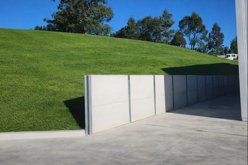 Concrete wall separating a grassy hill from a paved area, with trees and blue sky in the background.