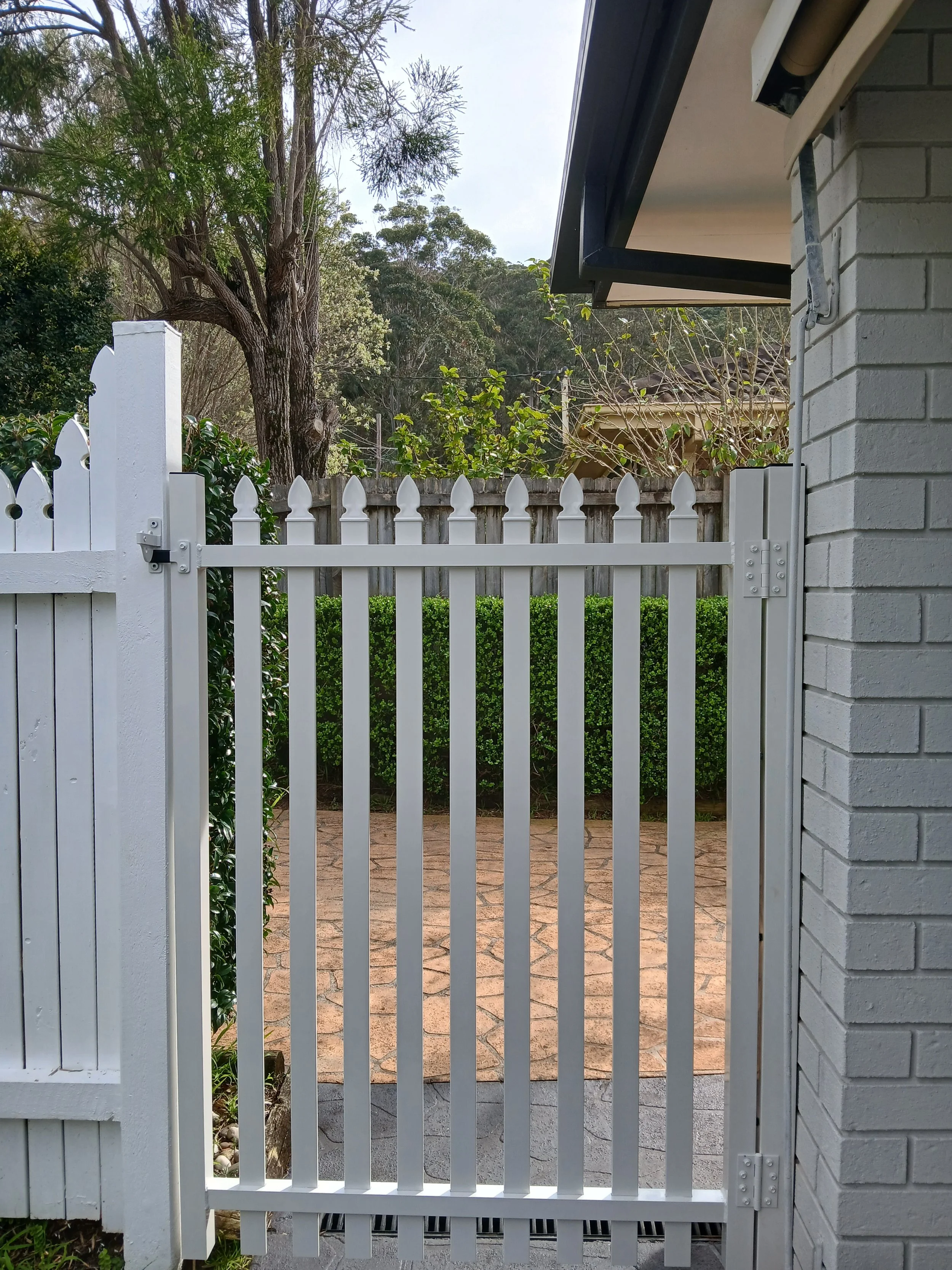 A white wooden gate with a latch, leading to a brick-paved backyard area, with green bushes, a tall tree, and a wooden fence in the background.