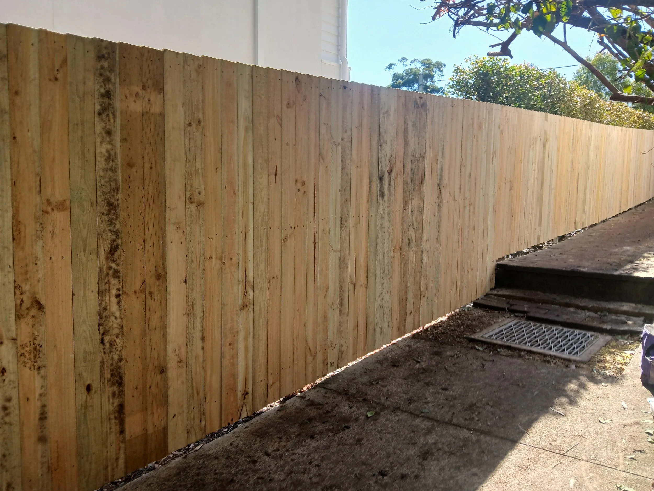 Newly installed wooden fence along sidewalk with steps and street drain visible, trees in background, sunny weather.