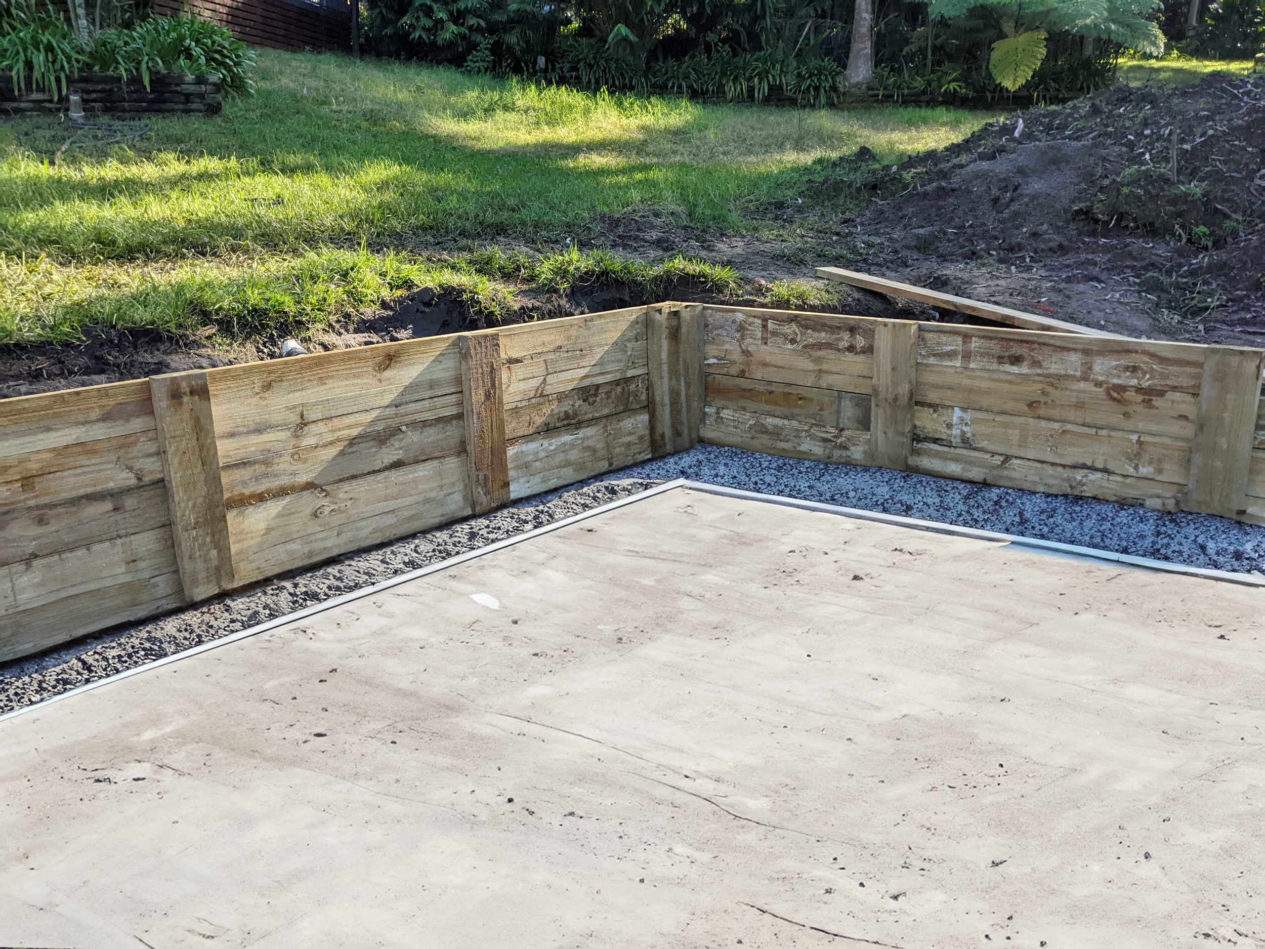 A wooden garden bed frame being constructed on a concrete surface, with grass and garden greenery in the background.