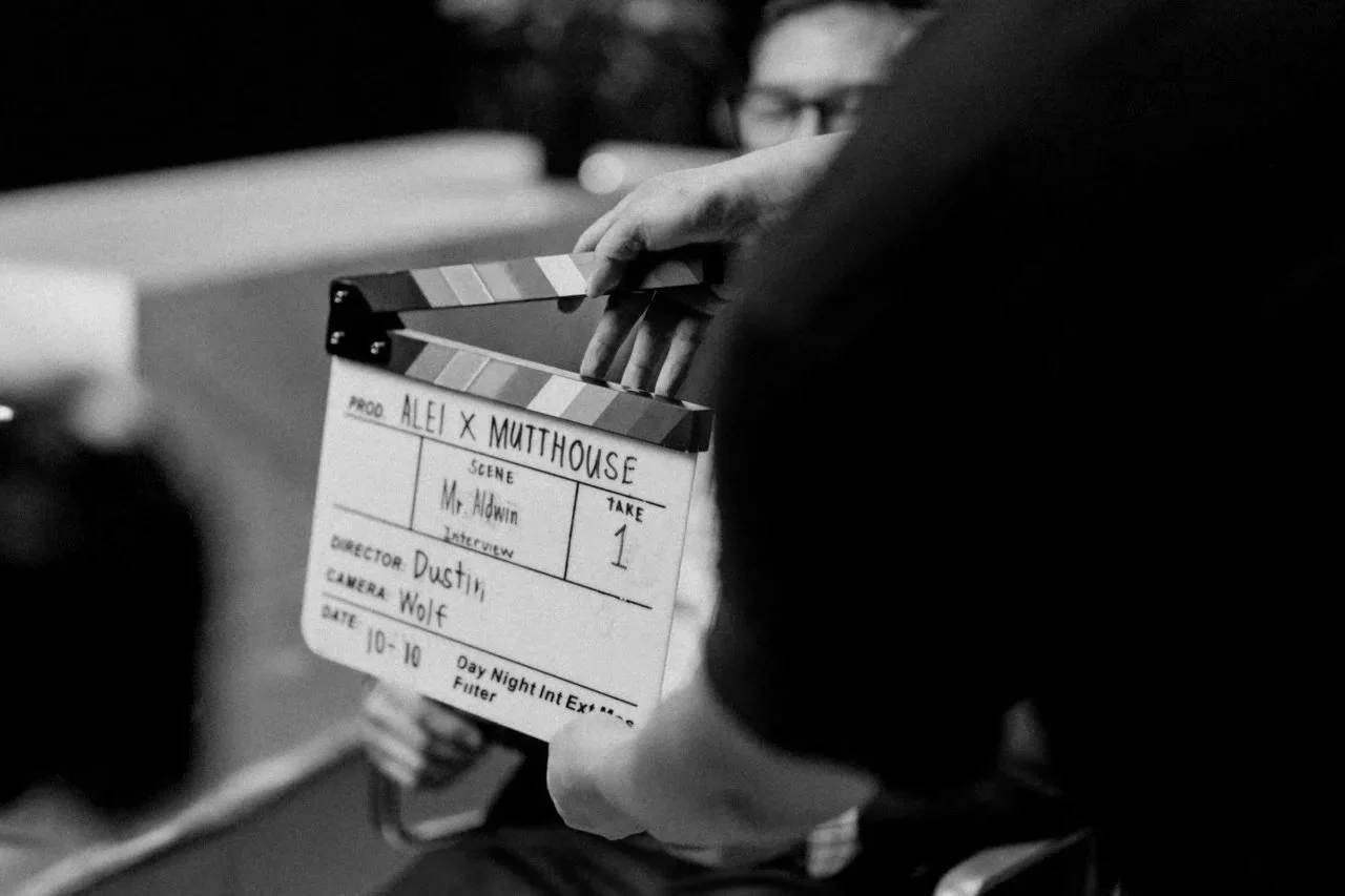 Close-up of a film slate or clapperboard held by a person, with another person blurred in the background. The slate has handwritten details including production title, scene, take, director, camera, and date.