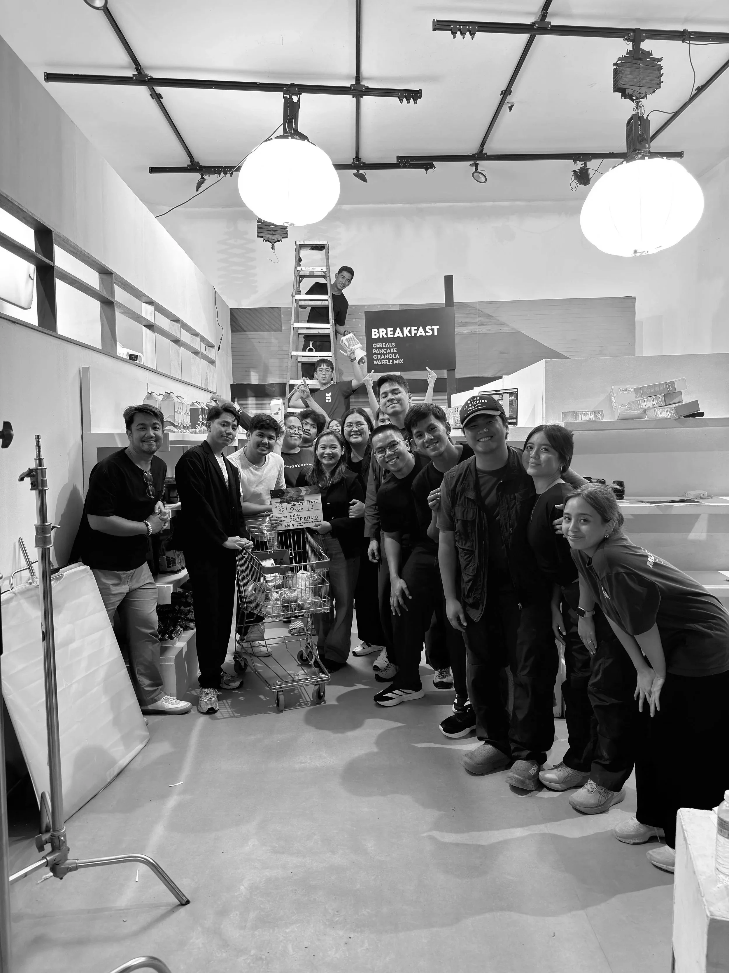 Group of people posing for a photo inside a store, with a shopping cart filled with groceries in front of them, and a person on a ladder in the background near a sign that says 'BREAKFAST'.
