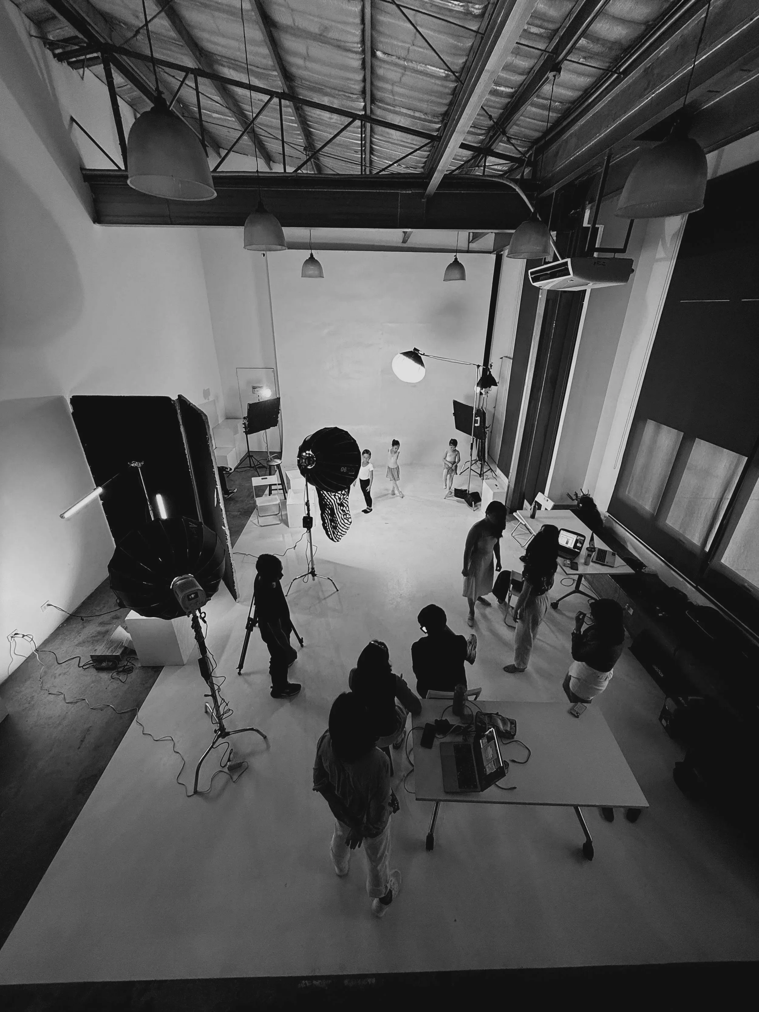 A woman is taking a photo of a young girl who is seated on a stool in a photo studio with a dark backdrop. The girl is smiling, wearing a checkered shirt, and the woman is facing her, holding a camera or phone.
