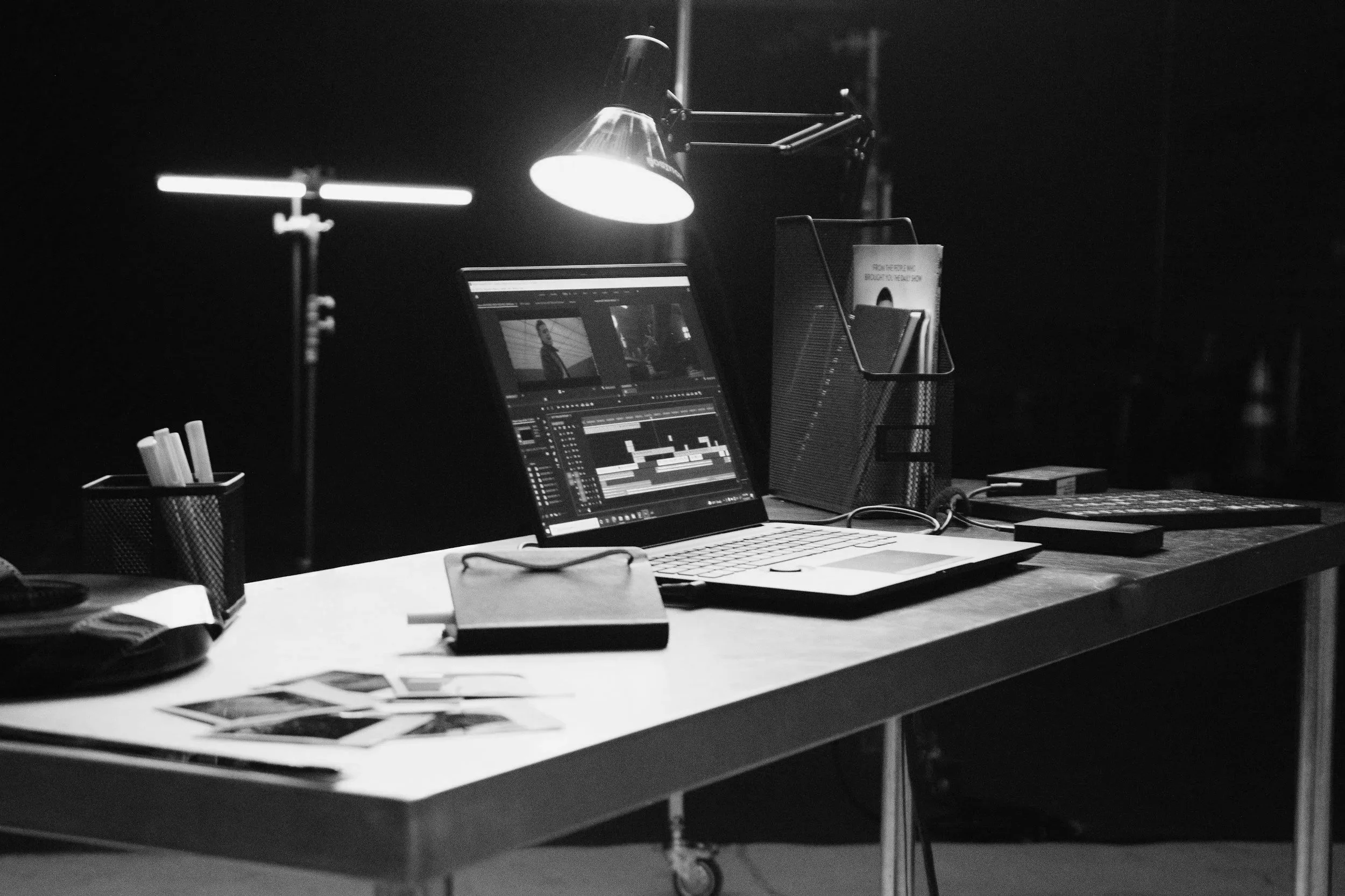 A workspace with a laptop on a white desk, displaying a video editing software, surrounded by photographs, a notebook, a pen holder, and other office supplies, with a lamp overhead illuminating the area.