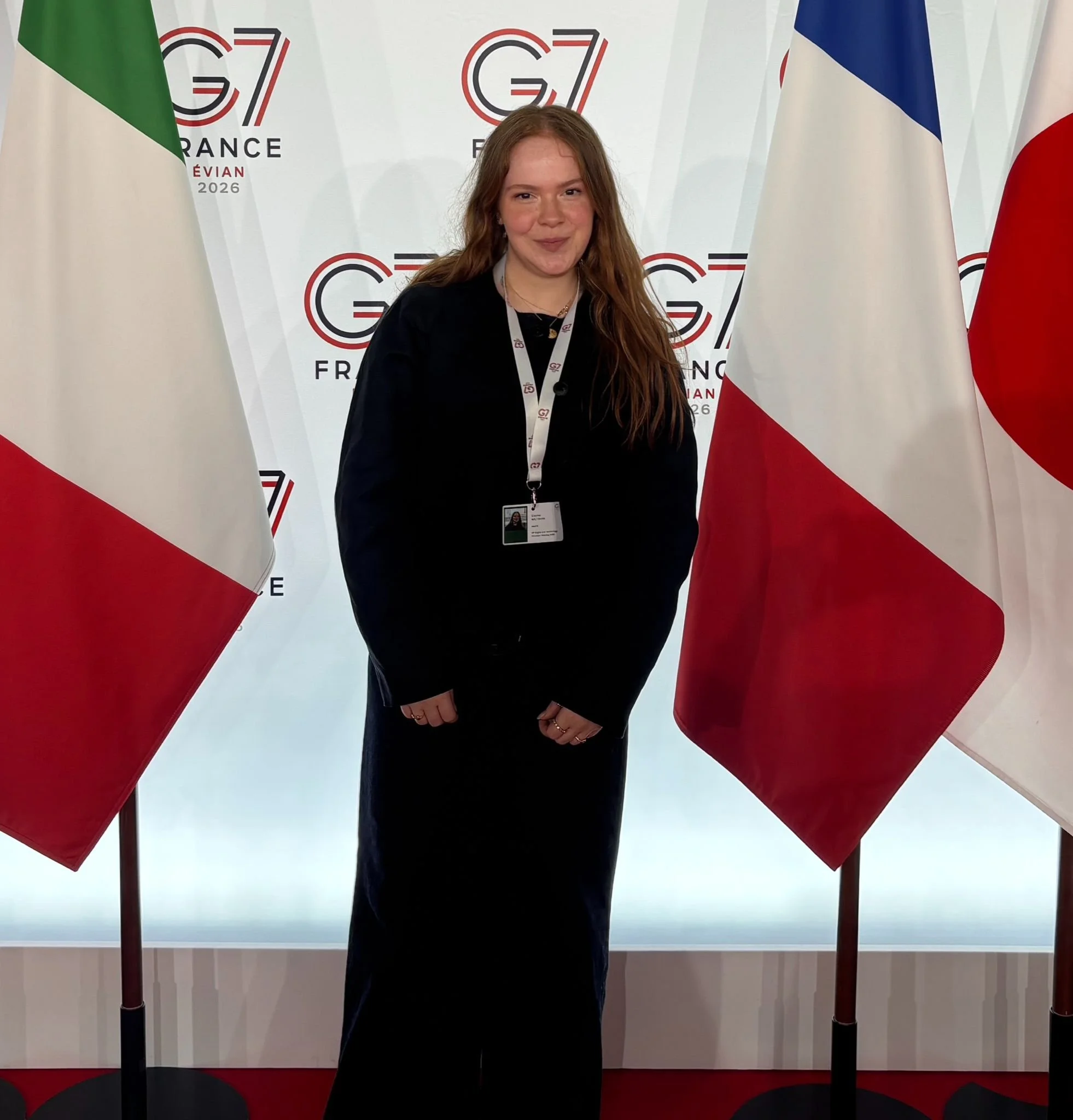 A young woman with long red hair smiling while standing in front of European and Canadian flags.