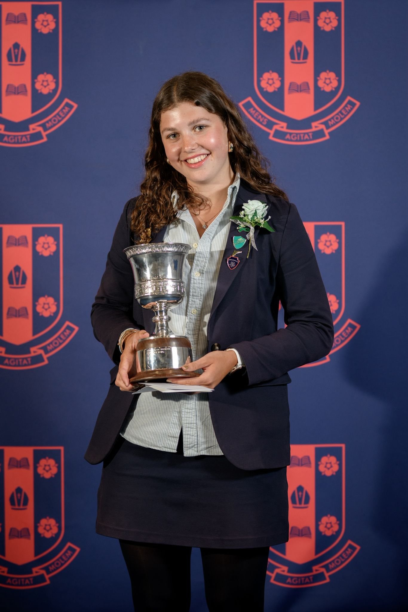 Young woman with curly hair wearing a blazer with a white flower boutonniere, holding a silver trophy, standing in front of a backdrop with a crest and the Latin motto 'Mens Agitat Molem'.
