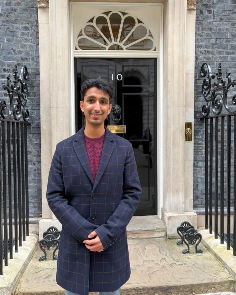 A young man standing in front of a black door with the number 10, surrounded by a stone frame and iron gates, on a gray stone porch. He is wearing a dark plaid coat and a maroon sweater, smiling at the camera.