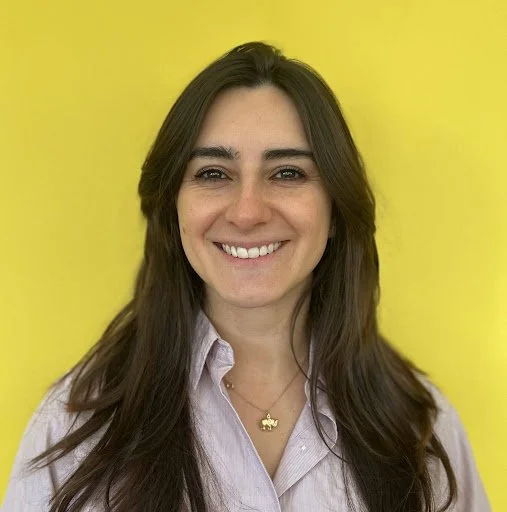 A woman with long brown hair smiling in front of a yellow background, wearing a light purple button-up shirt and a gold necklace.