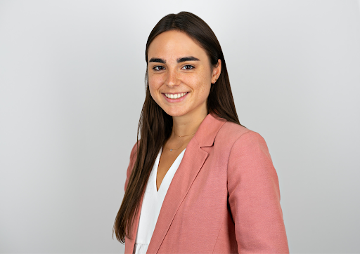 A woman with long brown hair wearing a pink blazer and white blouse, smiling against a plain light gray background.