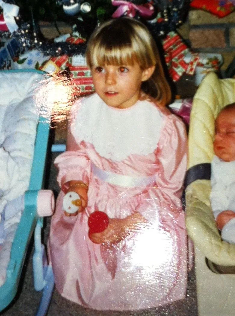 A young girl with light brown hair and bangs sitting in front of a Christmas tree, wearing a pink dress with white lace. Next to her, there is a baby in a yellow outfit. The scene appears to be a family holiday celebration with wrapped presents in the background.