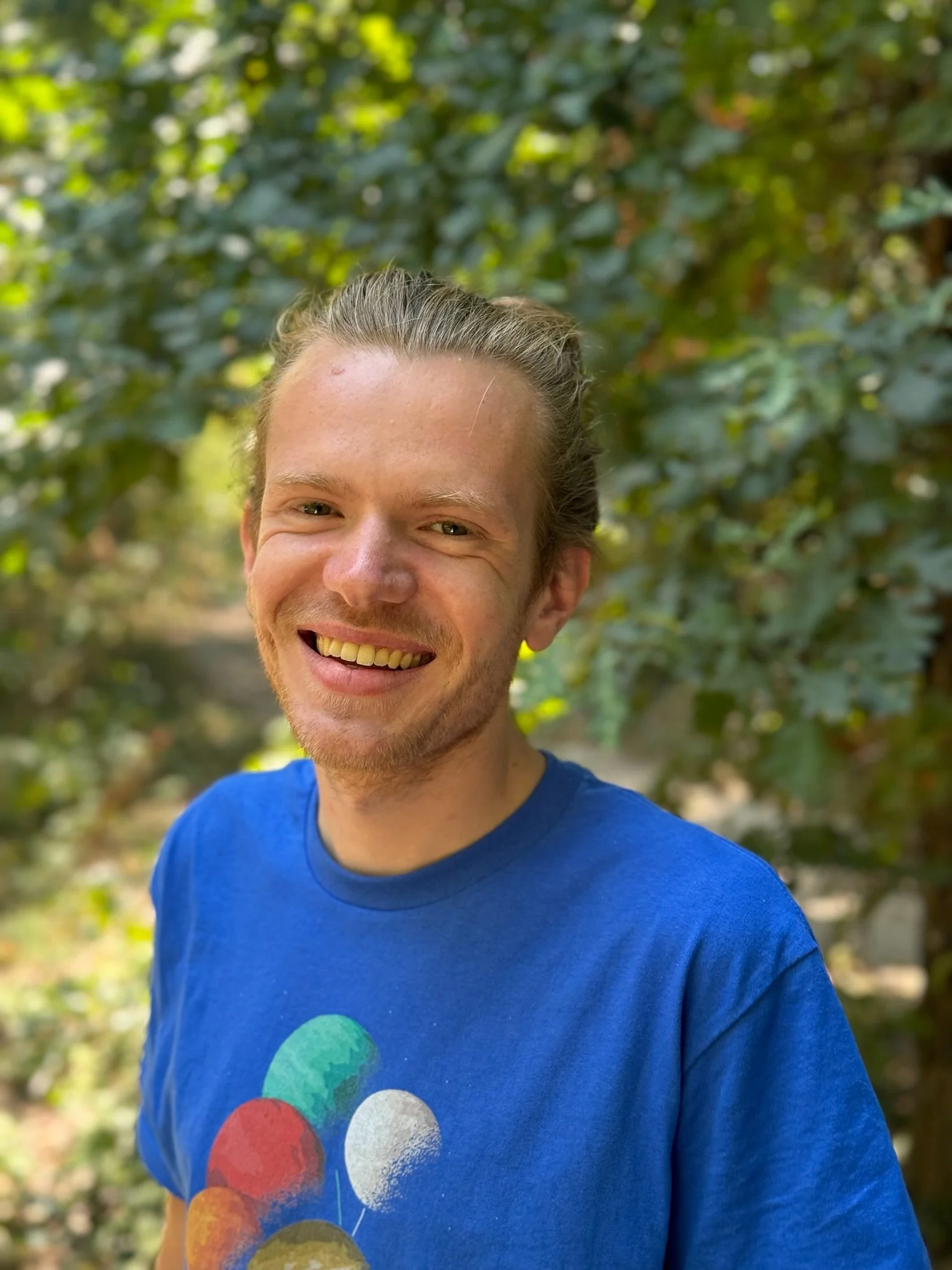 A smiling young man with light skin, blonde hair tied back, wearing a blue t-shirt with colorful dots, standing outdoors with green foliage in the background.