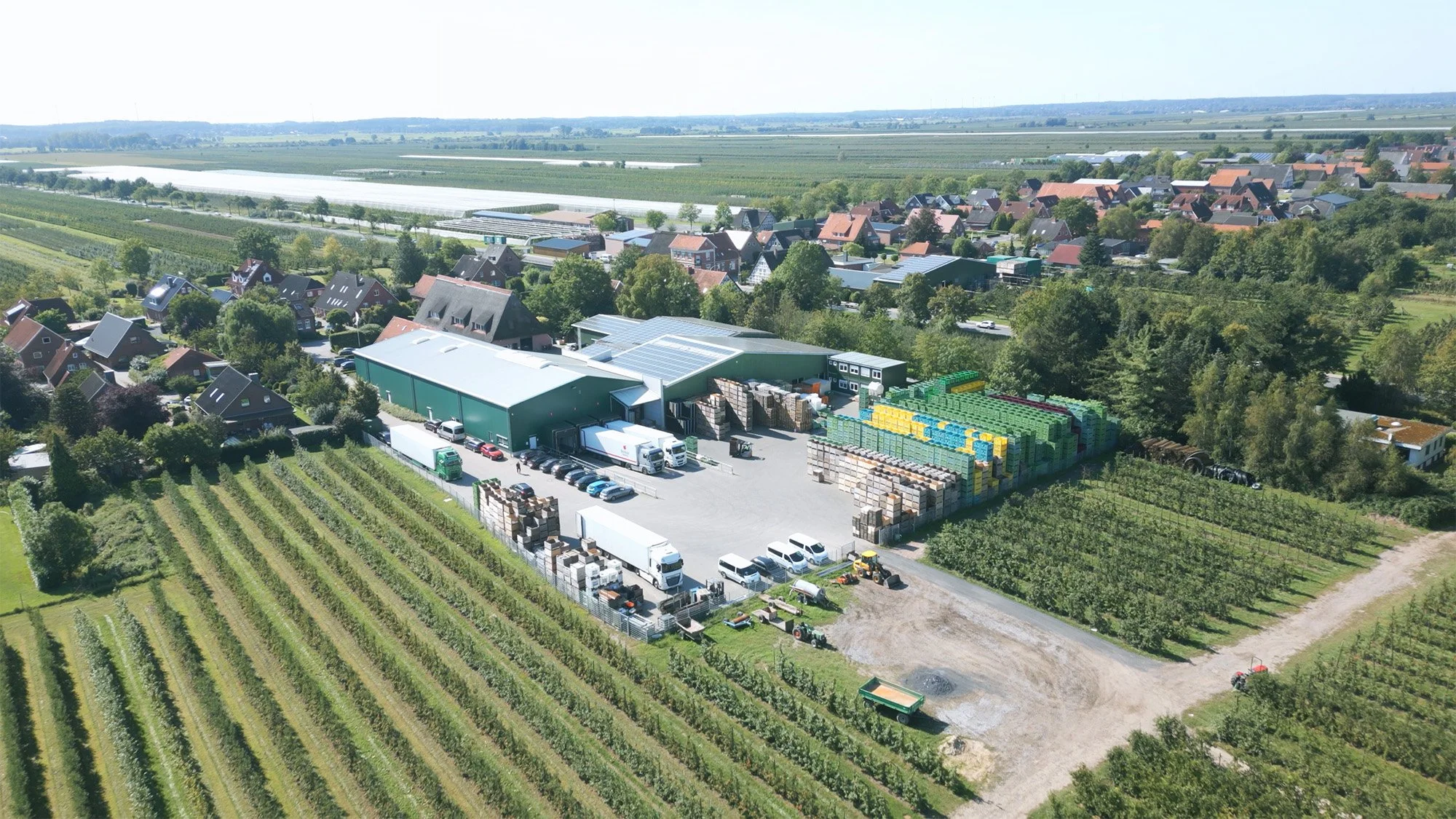 Luftaufnahme einer landwirtschaftlichen Betrieb mit Lagerhalle, landwirtschaftlichen Fahrzeugen, Getränkeverpackungen und Feldern in einem kleinen Dorf.