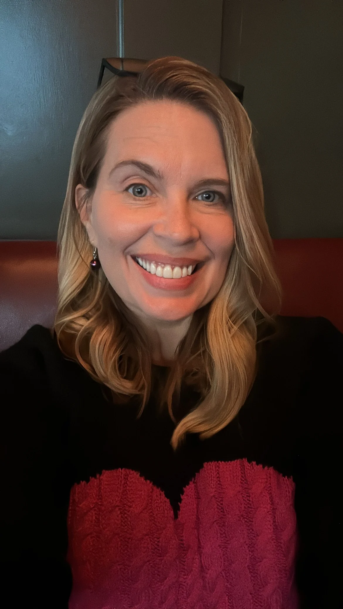 A woman with long blonde hair and blue eyes smiling, wearing black and red clothing, sitting indoors with a dark wall and a lamp in the background.