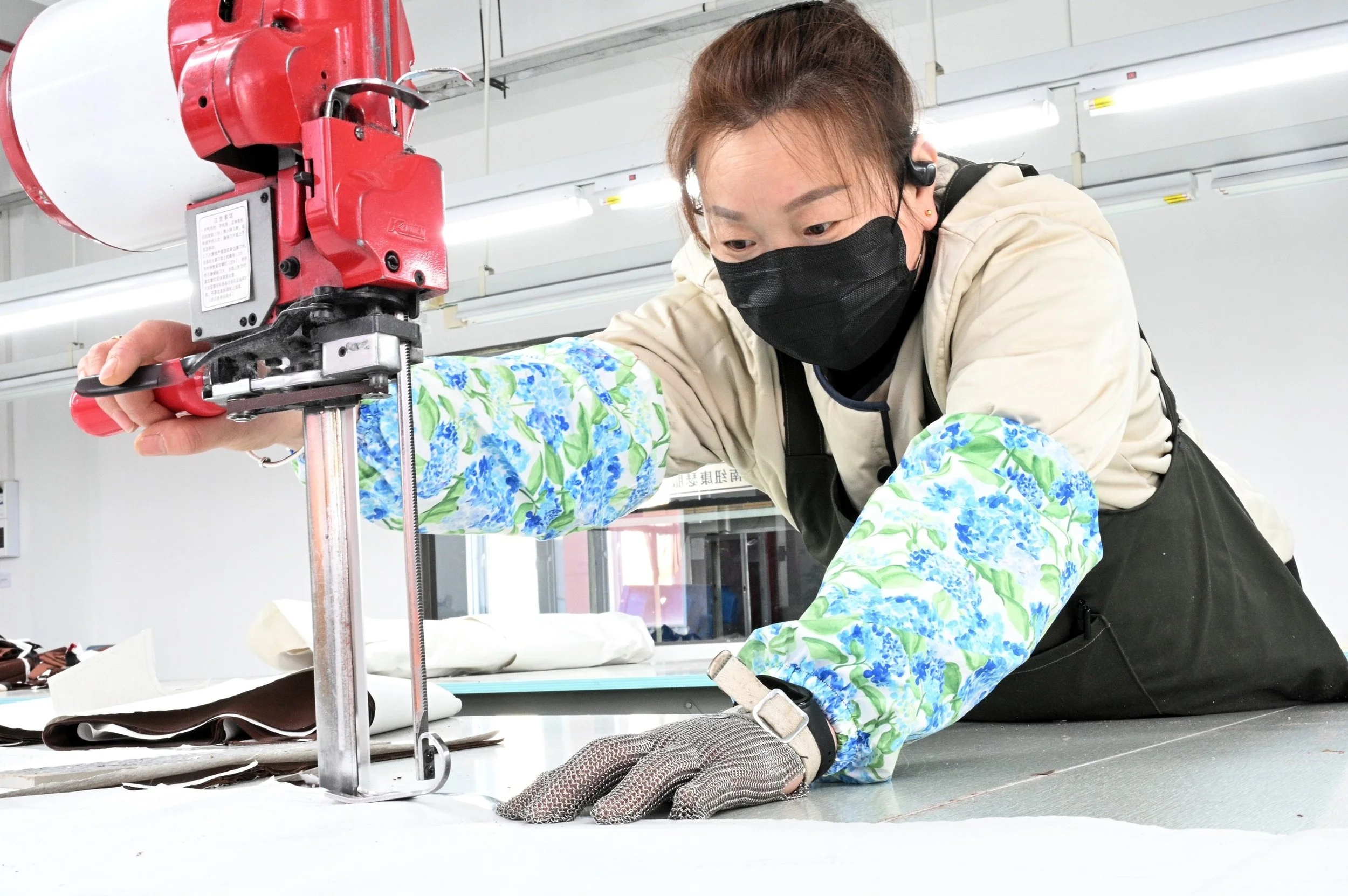 A woman wearing a black face mask, gloves, and a white jacket with floral sleeves is working with a red industrial fabric cutting machine on a large piece of fabric on a work table.
