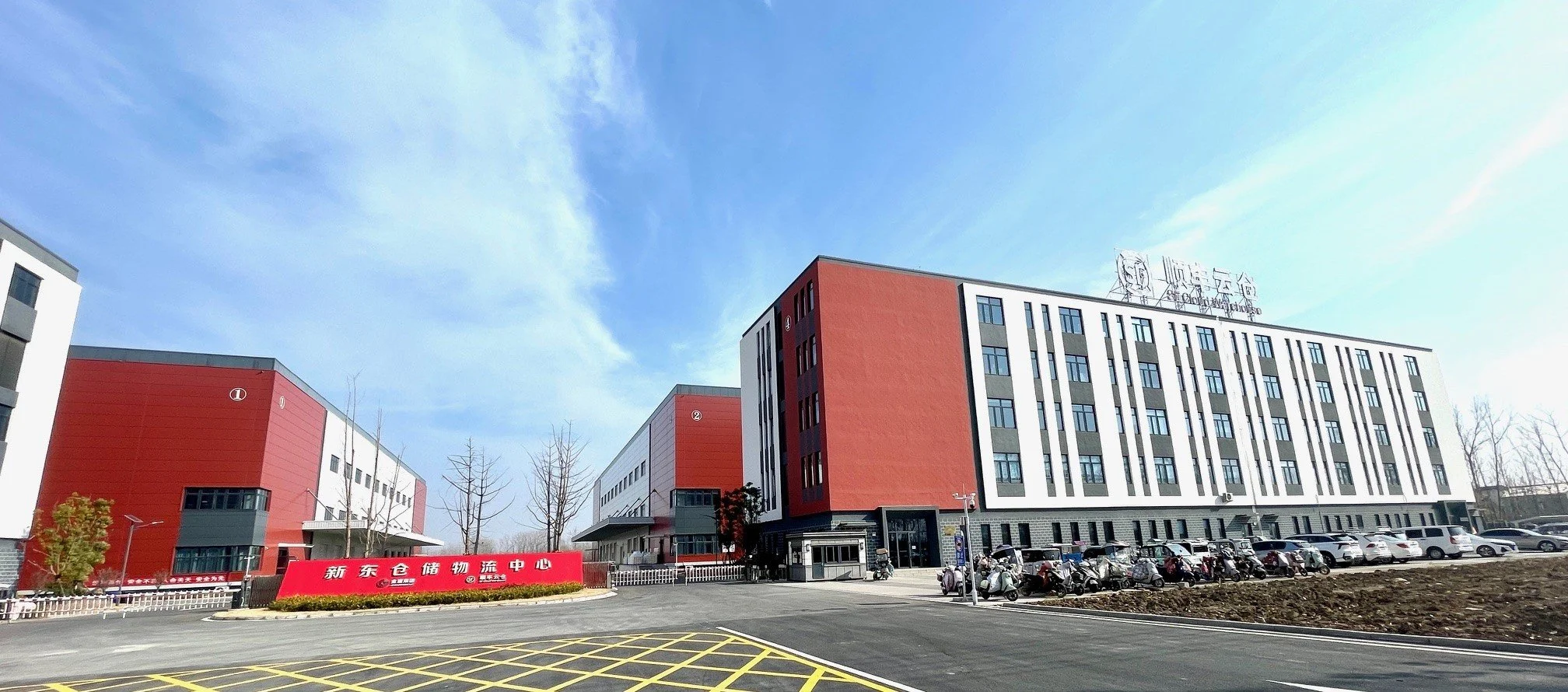 A modern logistics center building with a red and white exterior, parked motorcycles and cars, and a clear blue sky.