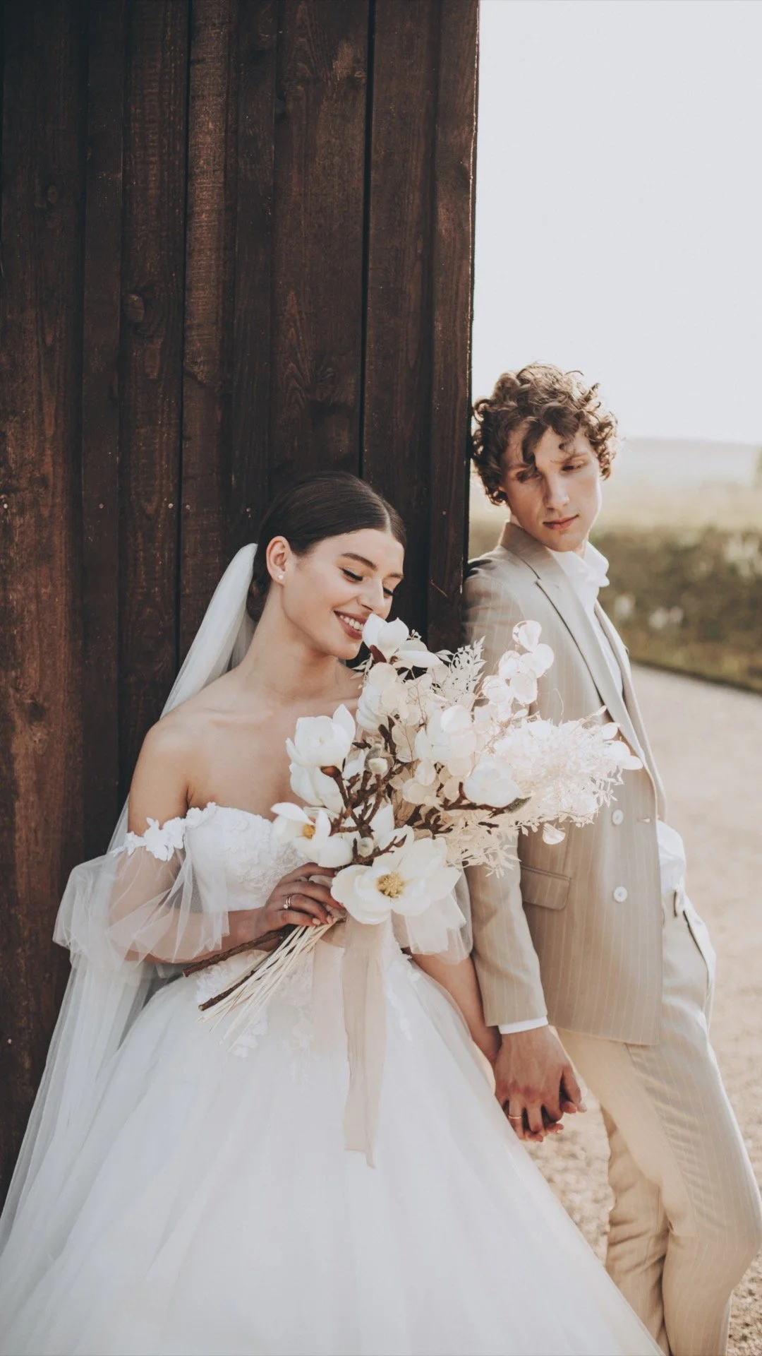 Un couple de mariés en costume blanc et beige, tenant la main, devant un mur en bois, avec la mariée tenant un bouquet blanc, souriant et regardant le bouquet, l'autre regardant le bouquet