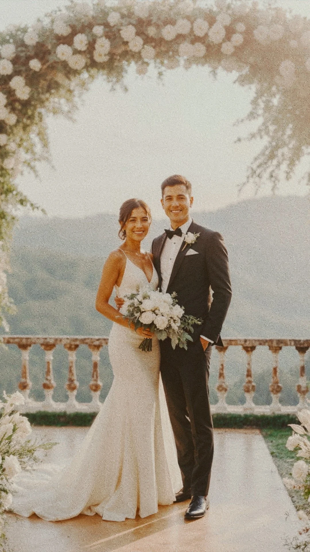 Un couple de mariés souriants posant ensemble lors de leur mariage, entourés de fleurs blanches, avec un décor en plein air et une vue sur des montagnes en arrière-plan.