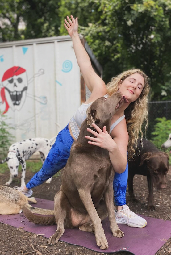 A woman in yoga attire, performing a stretching pose on a mat outdoors, surrounded by several dogs. One dog is affectionately licking her face. There's a mural of a pirate skull in the background.