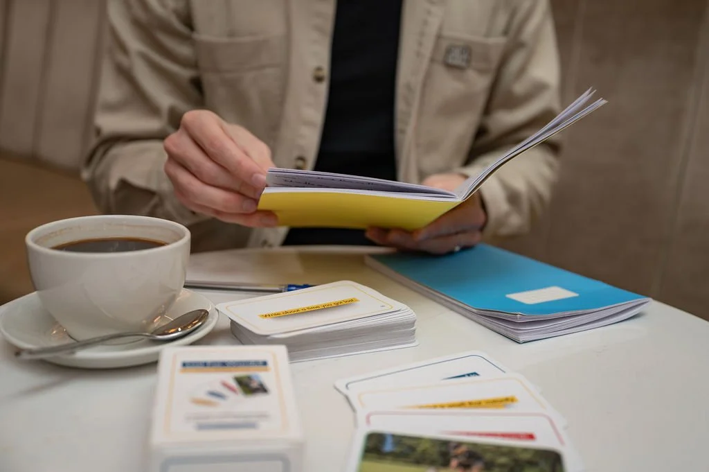 Person writing in yellow notebook with Lost For Words Creative Writing Prompt Cards on table