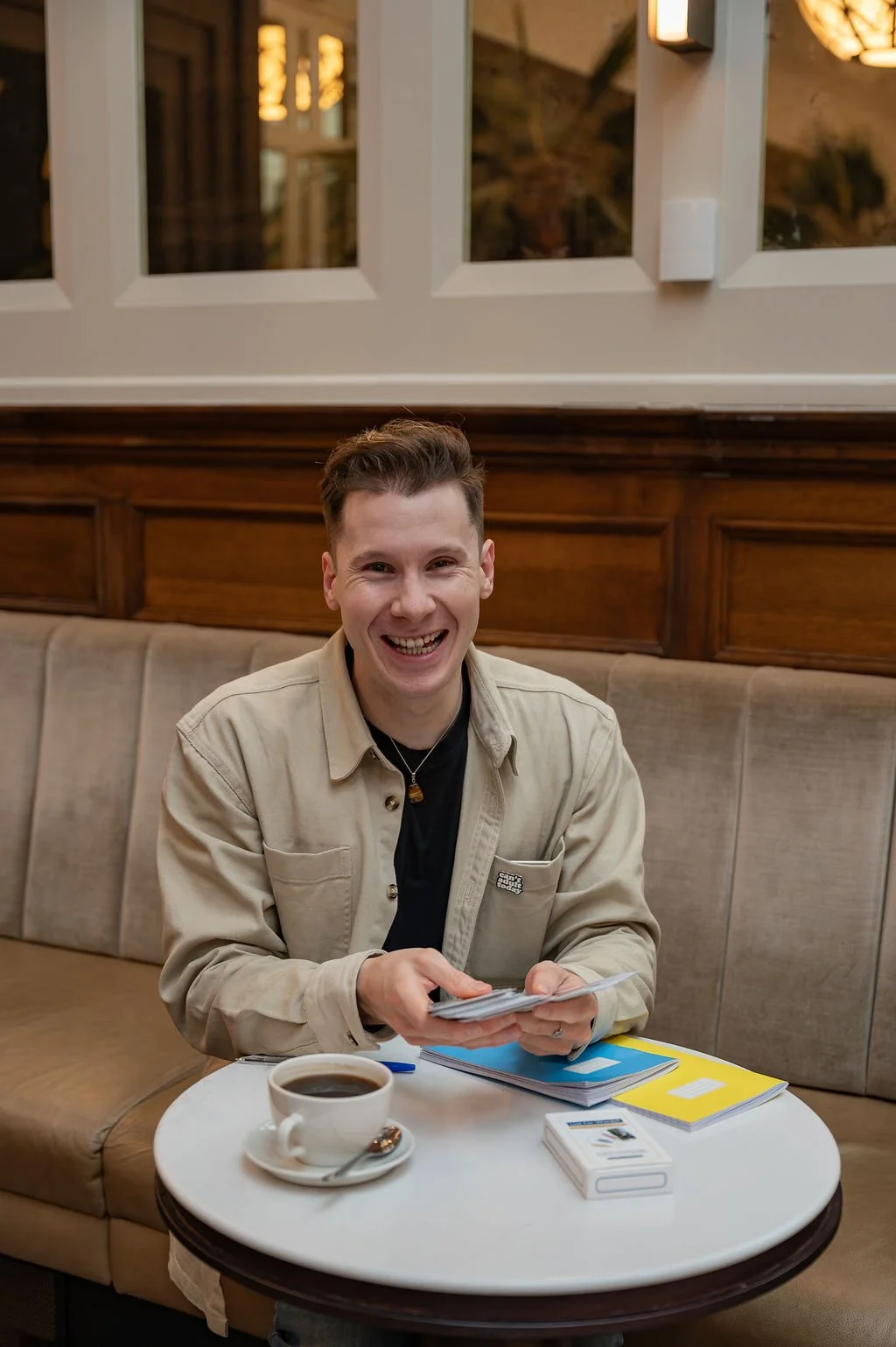 An image of writer and creative facilitator Liam Marshall. He is sat at a table with notebooks and a cup of coffee in front of him. He is looking into the camera and laughing, holding a pack of creative writing prompt cards.