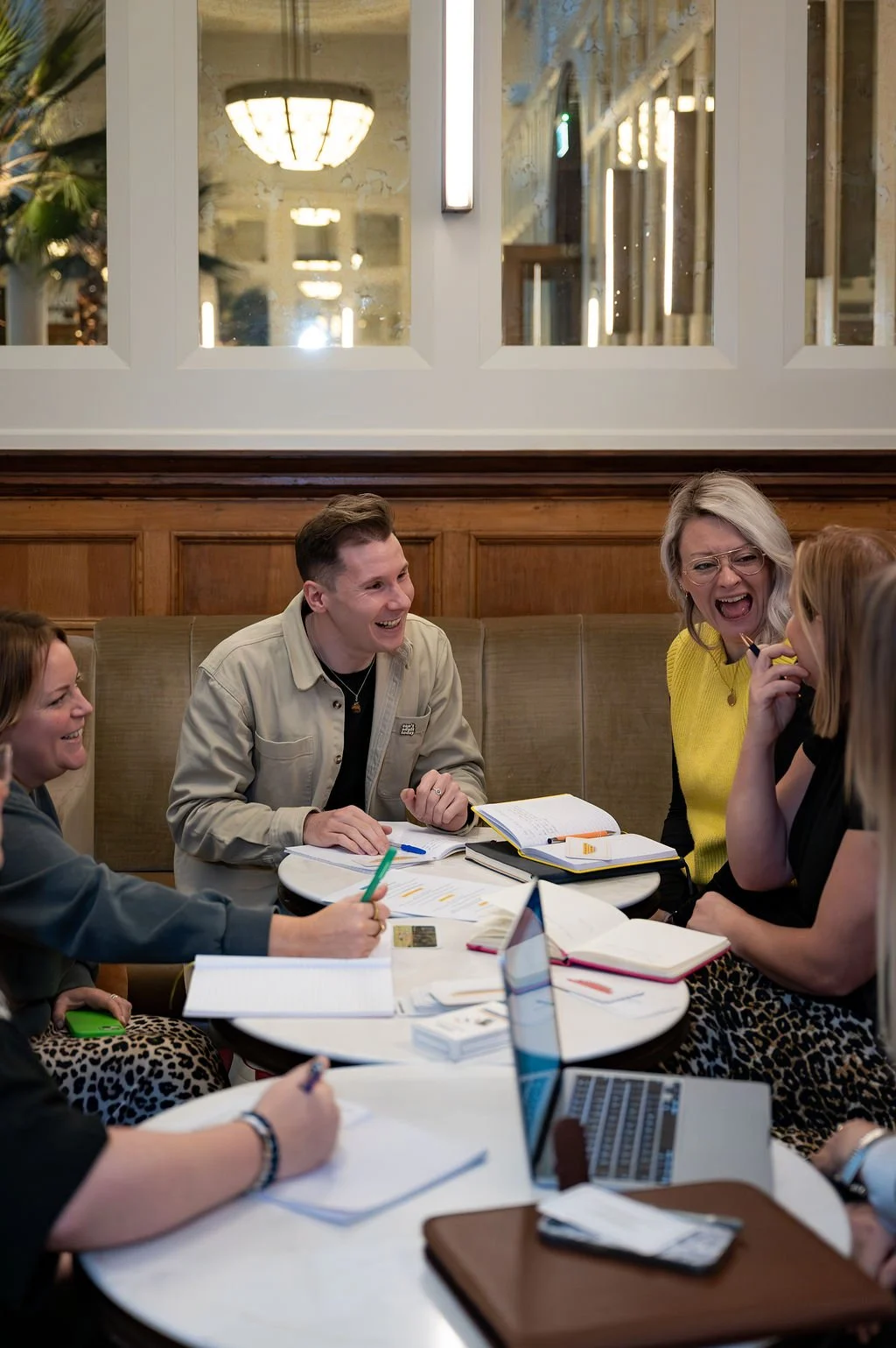 Liam Marshall facilitating a creative writing workshop, laughing with participants around a table covered in notebooks and writing materials.