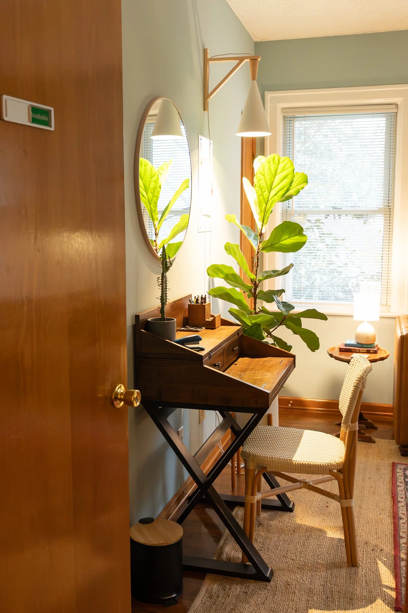 Cozy room with a vintage writing desk, a woven chair, a large fiddle-leaf fig plant near a window, a small side table with a lamp, and a wall mirror reflecting the plant.