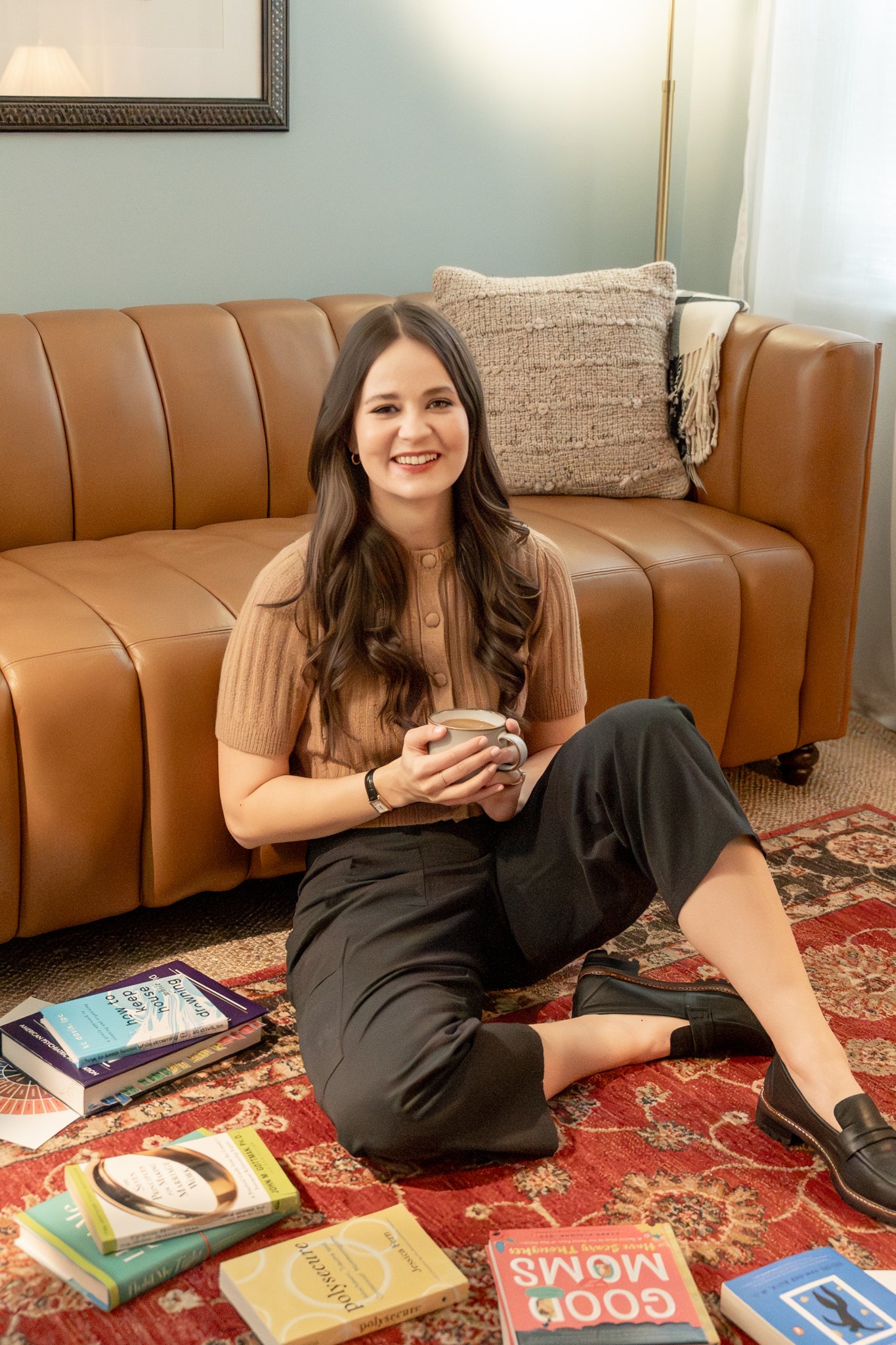 A young woman with long brown hair, wearing a tan short-sleeve top and black pants, sitting on a colorful area rug, holding a mug. Books are spread on the rug in front of her, and she is seated on the floor with one knee up. Behind her is a brown leather sofa with a textured cushion and a blanket, in a room with a wall mirror and a standing lamp.