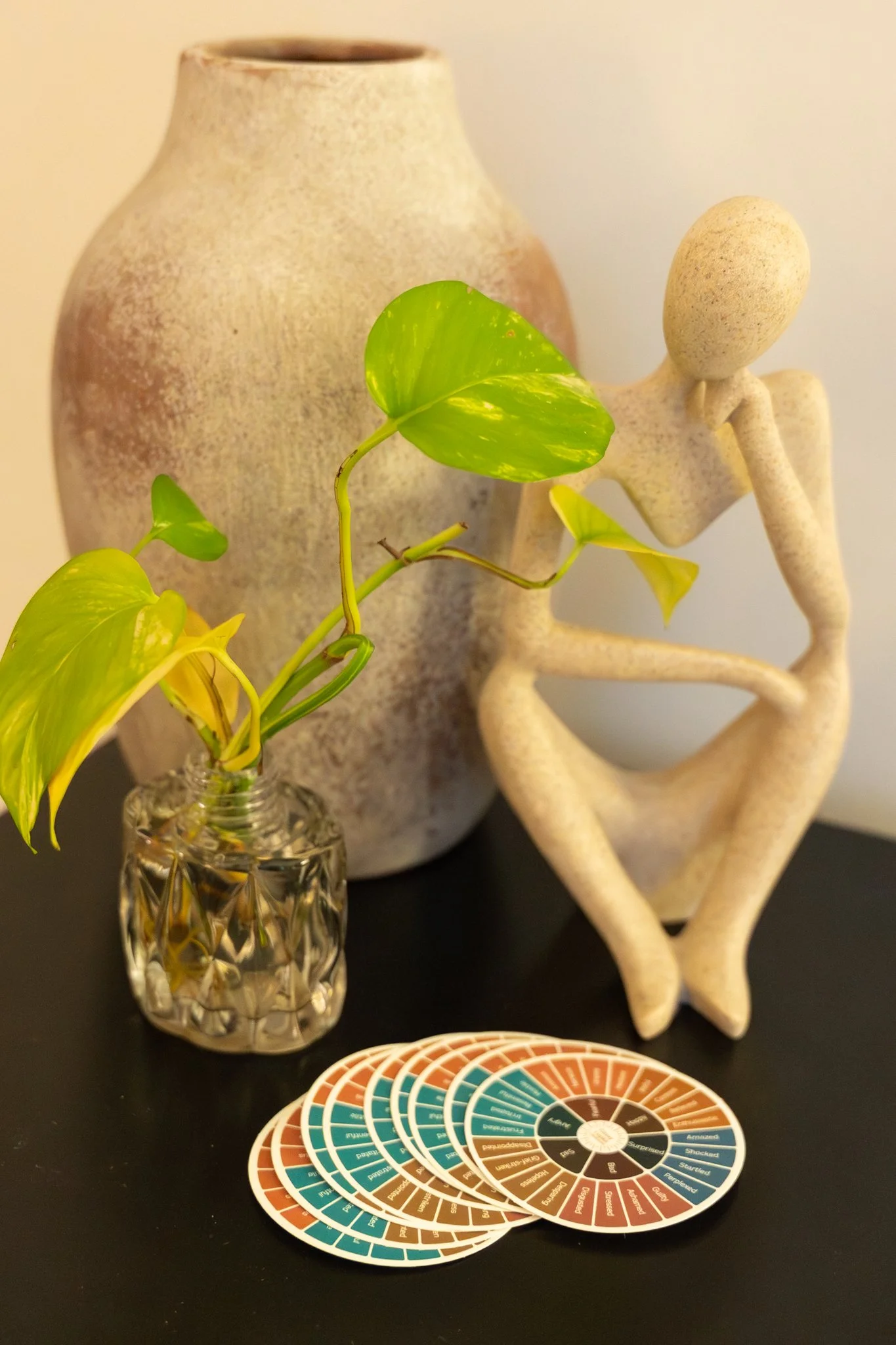 Decorative scene with a textured beige vase, a small glass vase with a green leafy plant, a abstract sculpture of a seated figure with knees drawn up and head resting on one arm, and a set of color swatches or fan charts with earthy tones.