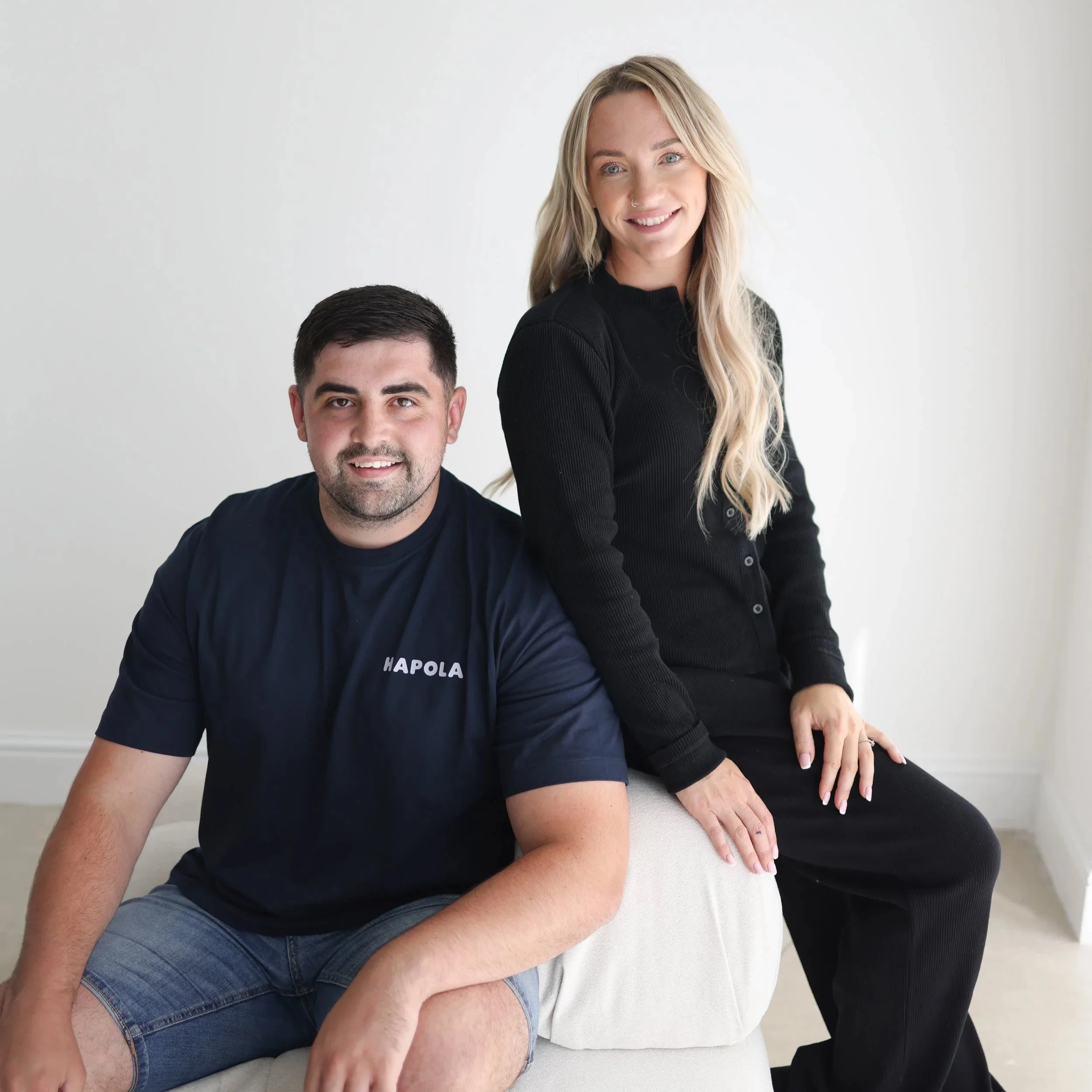 A man with dark hair and beard wearing a navy t-shirt and denim shorts, sitting on a beige sofa, next to a woman with long blonde hair wearing a black cardigan and black pants, smiling at the camera, in a minimalistic white room.