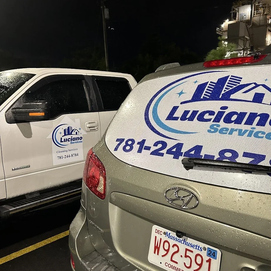 Two vehicles with "Luciano Services Cleaning Services" branding visible, parked side by side at night. The rear window of a Hyundai SUV displays a large logo and contact number, while a white pickup truck bears a similar logo and text on its door. A 