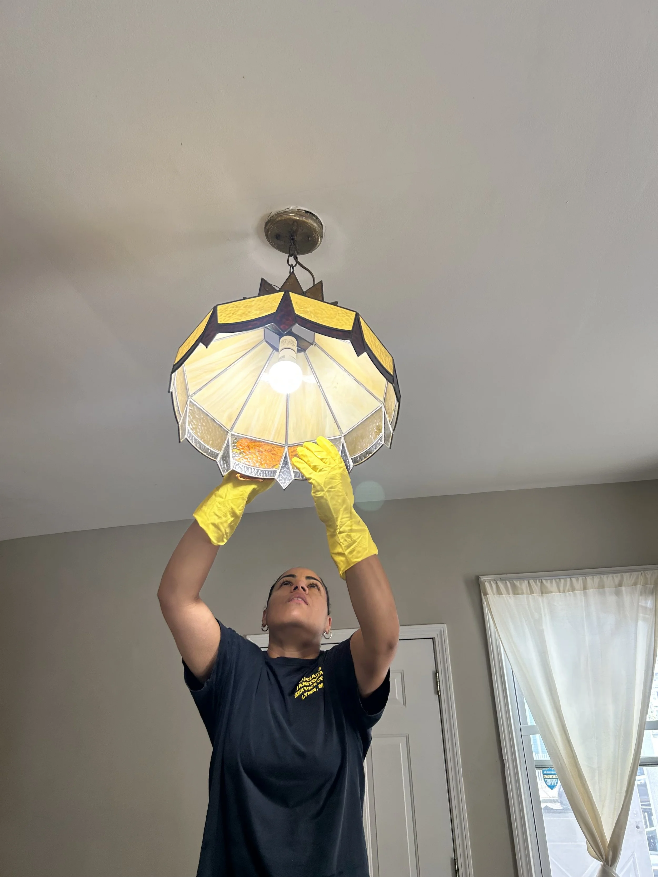A woman is standing indoors, reaching up with both yellow-gloved hands to adjust or install a stained glass ceiling pendant light fixture. She is wearing a black t-shirt and has her hair pulled back. The room has light-colored walls, a door, and a wi