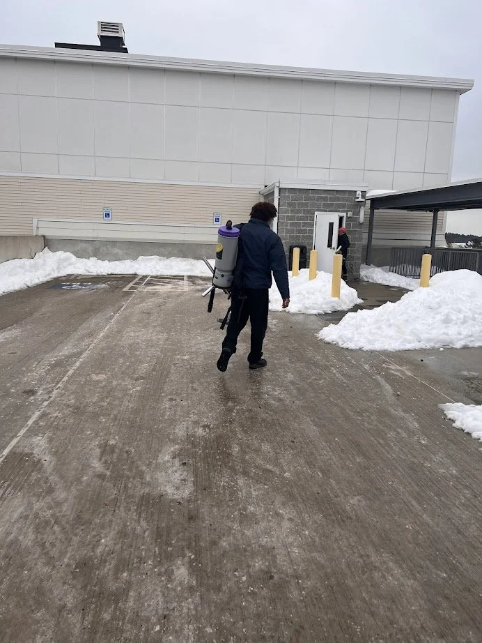 Person wearing a backpack leaf blower on a snowy path near a building.