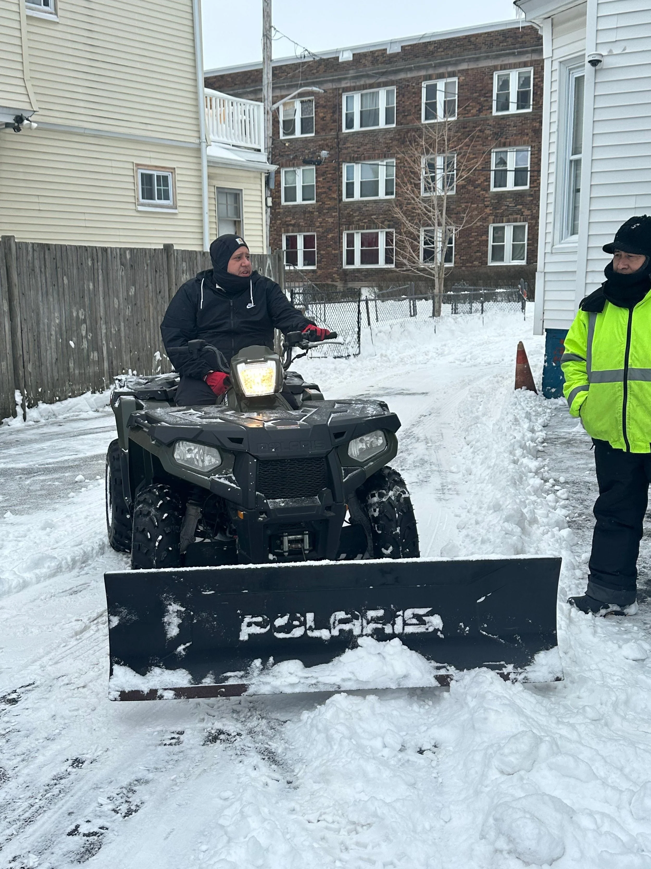 A man riding an ATV in a snowy residential backyard, with a snowplow blade attached, and another person standing nearby wearing a neon yellow safety jacket.