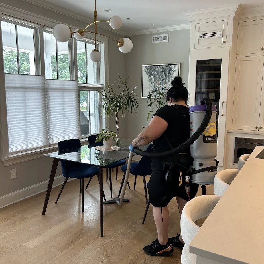 Person vacuuming a dining room with a backpack vacuum cleaner, featuring a glass table, blue chairs, and indoor plants.