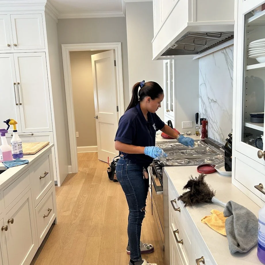 A woman wearing a navy blue uniform and blue gloves cleaning a stove in a bright, modern kitchen with white cabinets and light wood flooring.