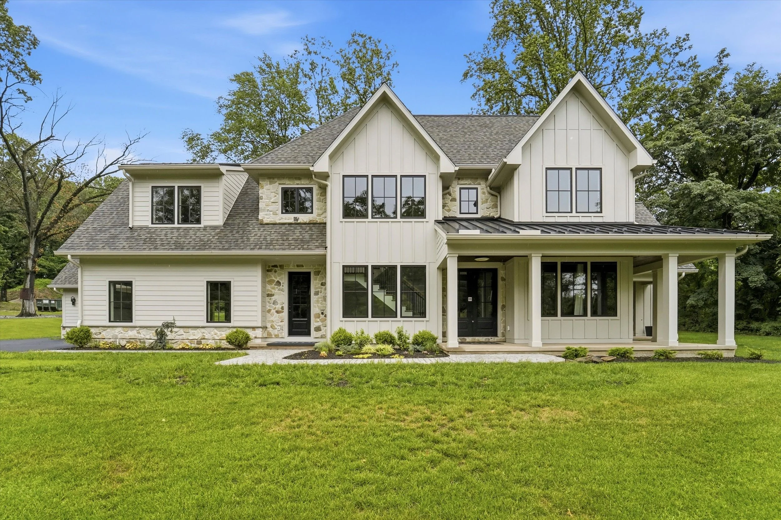 A large two-story house with a light exterior, black framed windows, stone accents, and a porch, surrounded by a green lawn and trees in the background under a blue sky.