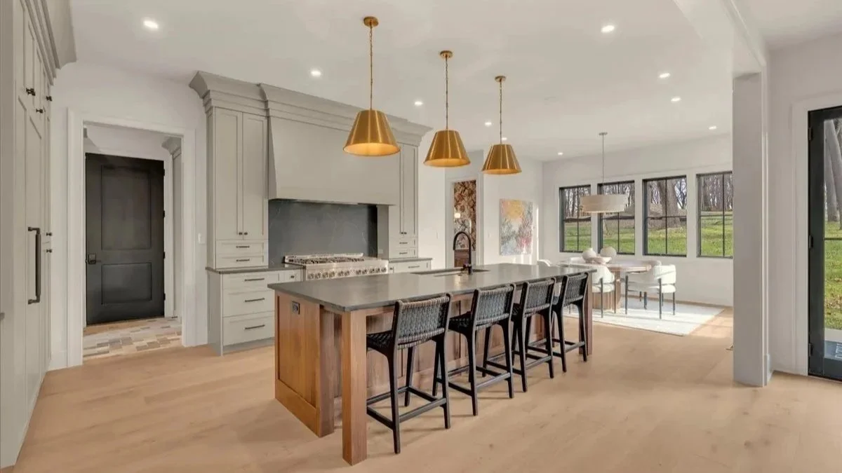 Open-concept kitchen and living area with light-colored walls and wood flooring, featuring a kitchen island with four black chairs, gold pendant lights, gray cabinets, and a large window view of a yard.