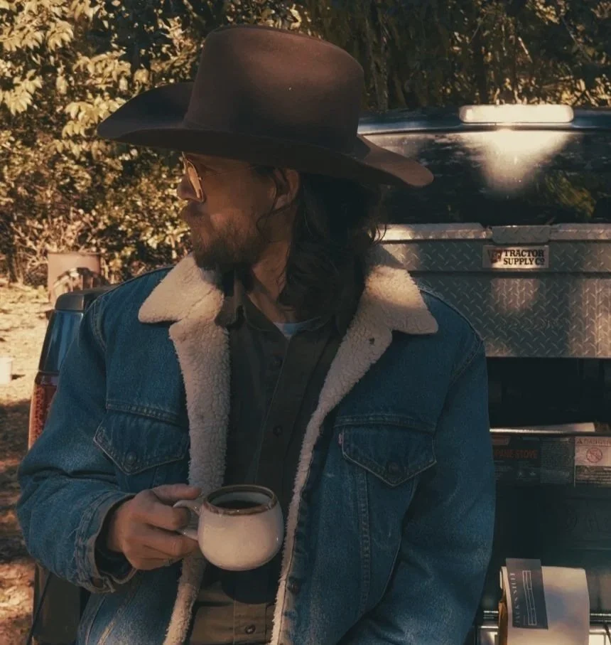A man with long dark hair and a beard wearing sunglasses, a cowboy hat, a denim jacket with a shear lining, and a dark shirt, holding a coffee mug, standing outdoors next to a truck with trees in the background.