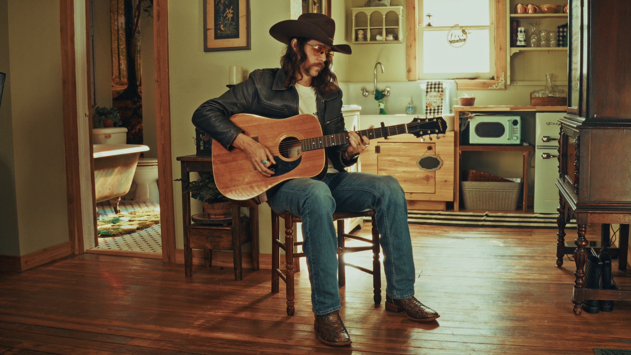 A man wearing a cowboy hat, glasses, leather jacket, and jeans playing an acoustic guitar while sitting on a wooden chair in a cozy kitchen.