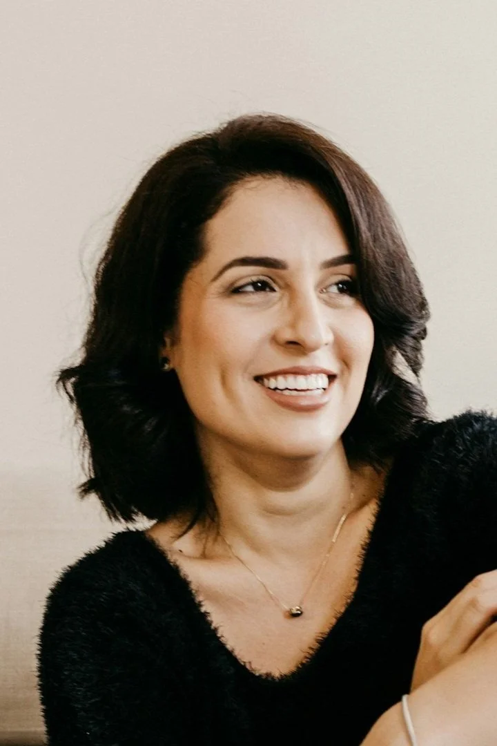 A woman with shoulder-length dark brown hair, smiling and wearing a black fuzzy sweater, a gold necklace, and earrings, standing against a plain beige wall.