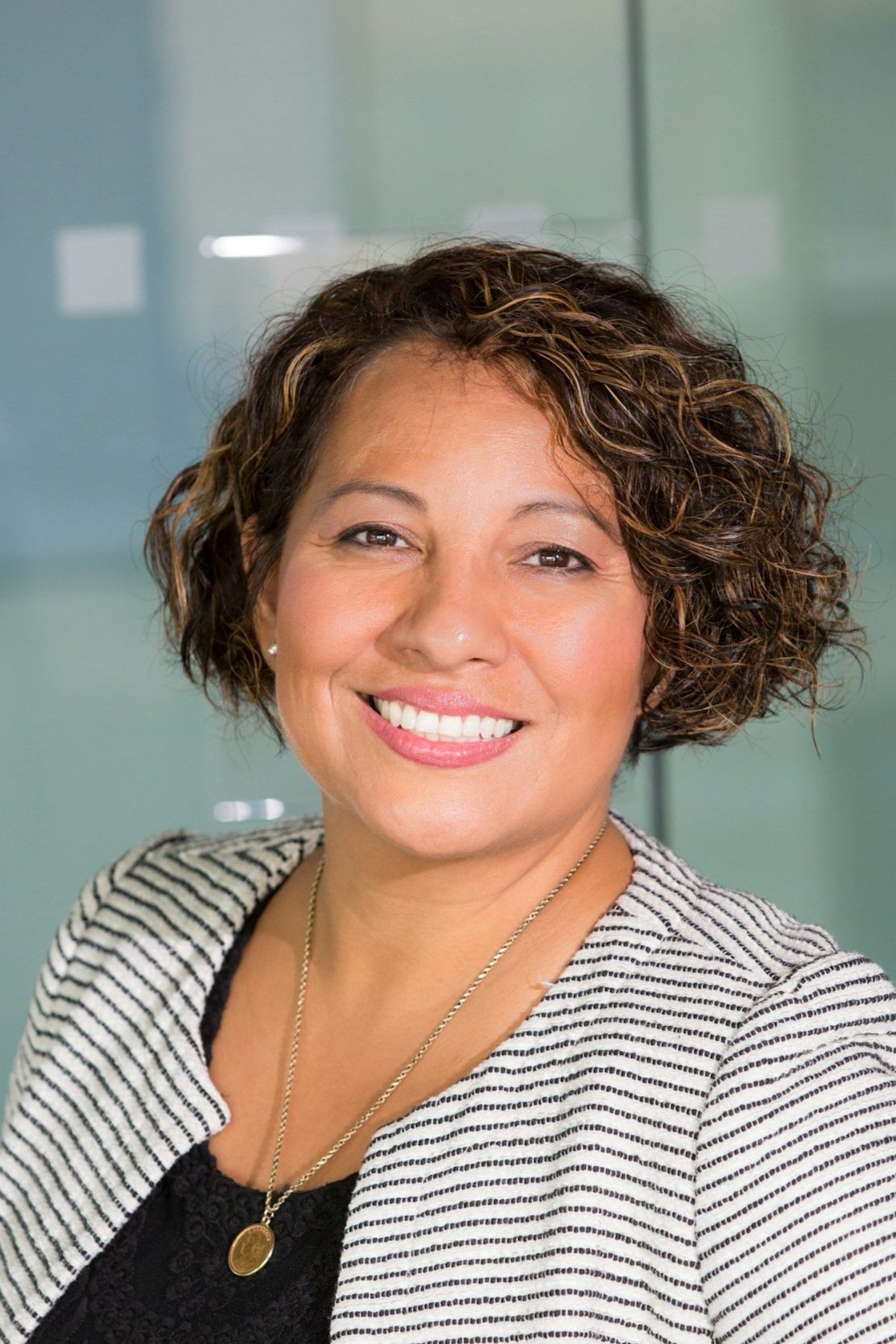 A woman with short, curly brown hair smiling, wearing a black top, a striped blazer, and a gold necklace, standing in front of a glass background.