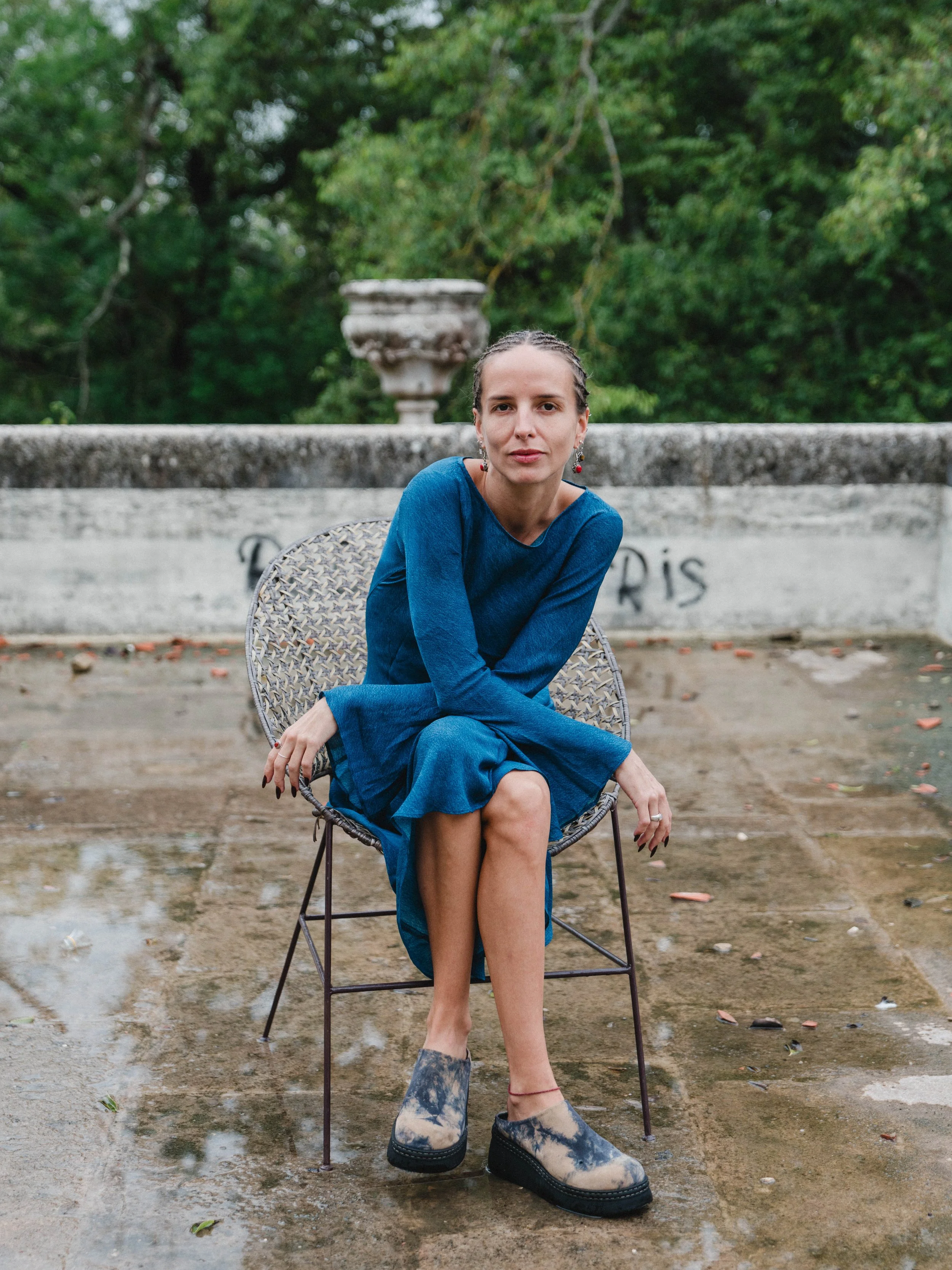 A woman in a blue dress sitting on a modern wire chair outdoors, with a concrete ledge and greenery in the background.