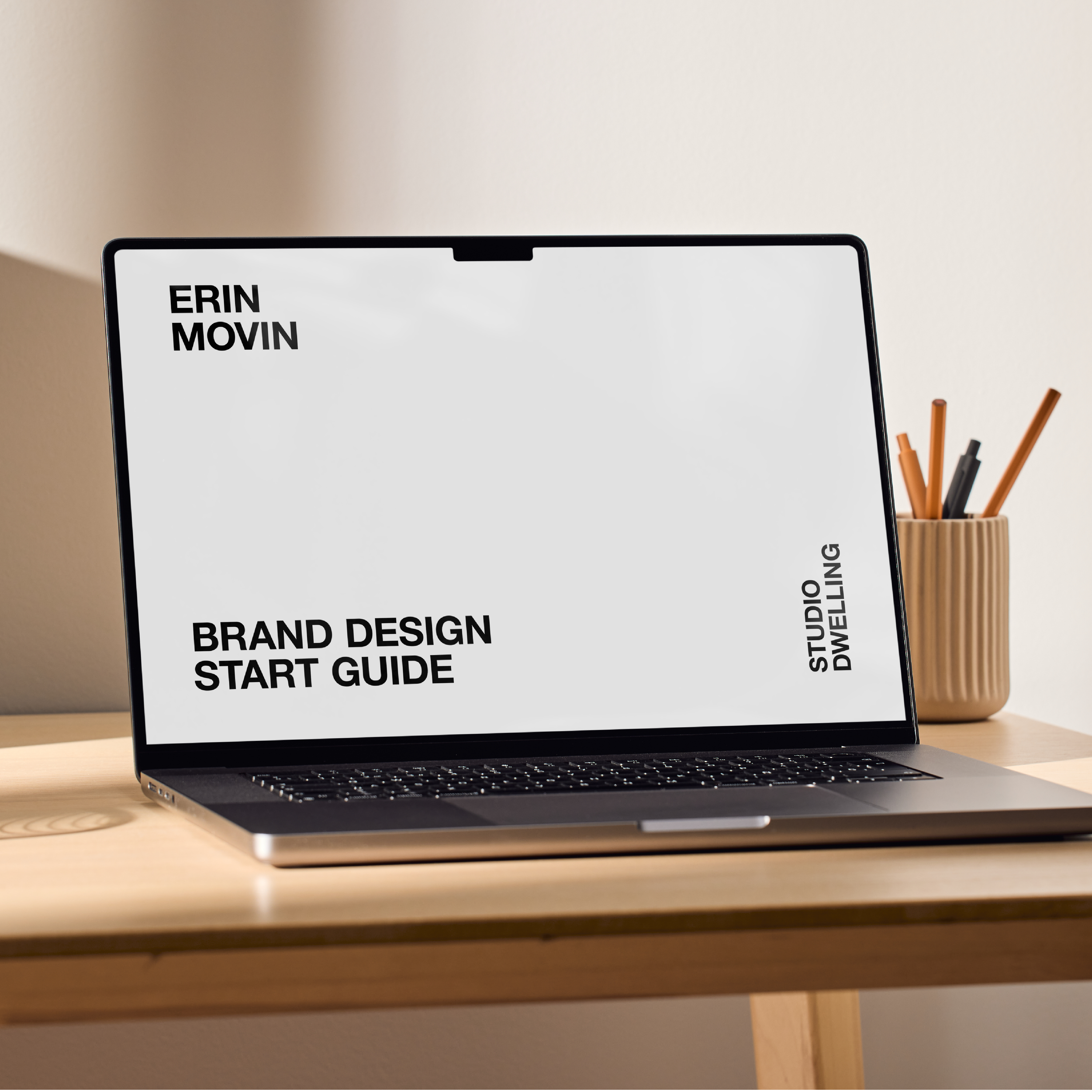 Laptop displaying "Brand Design Start Guide" on a wooden desk with a pencil holder in the background.
