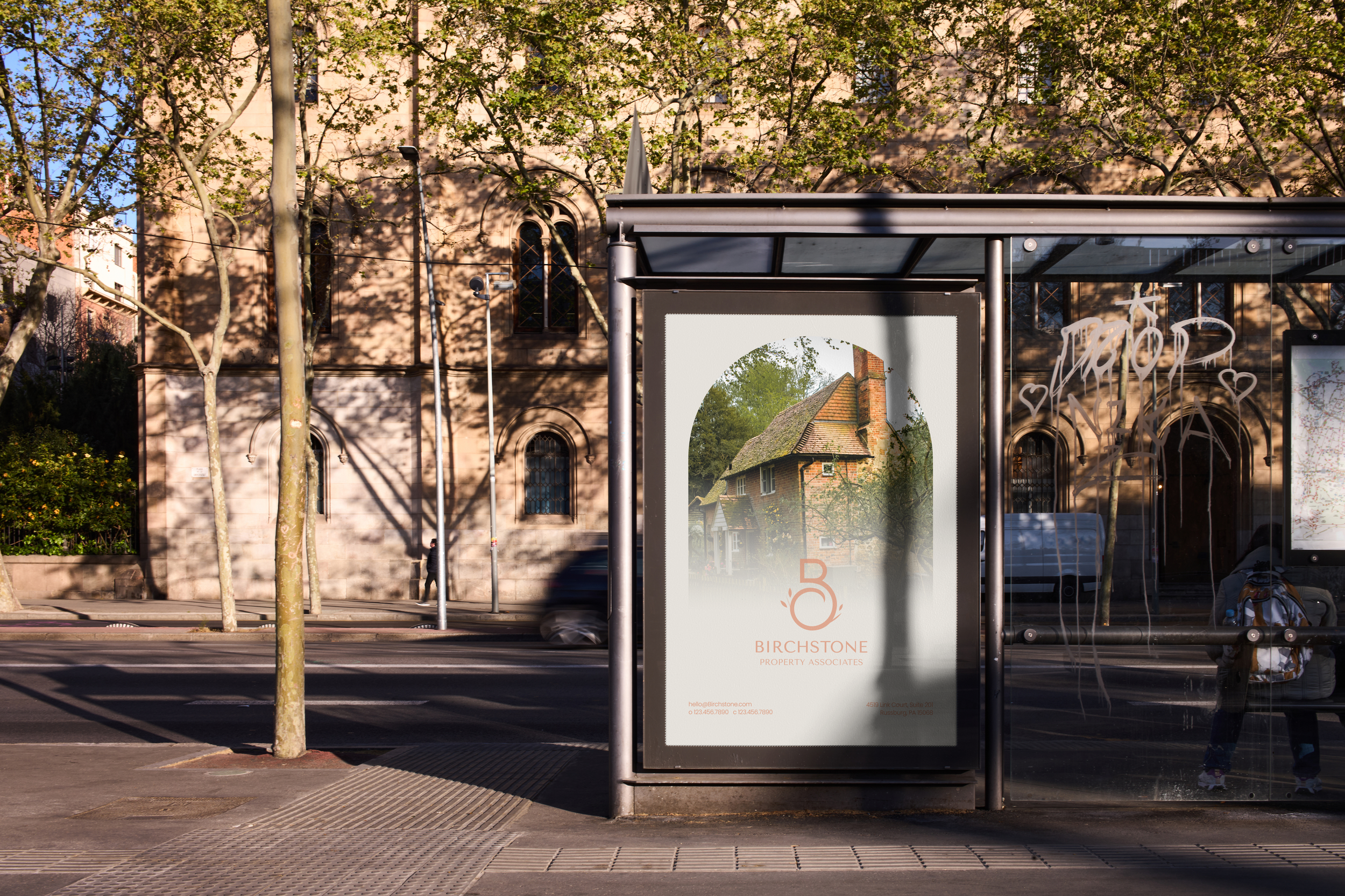 A bus stop with a Birchstone Property Associates advertisement and graffiti on the glass, located in front of an old stone building with arched windows.