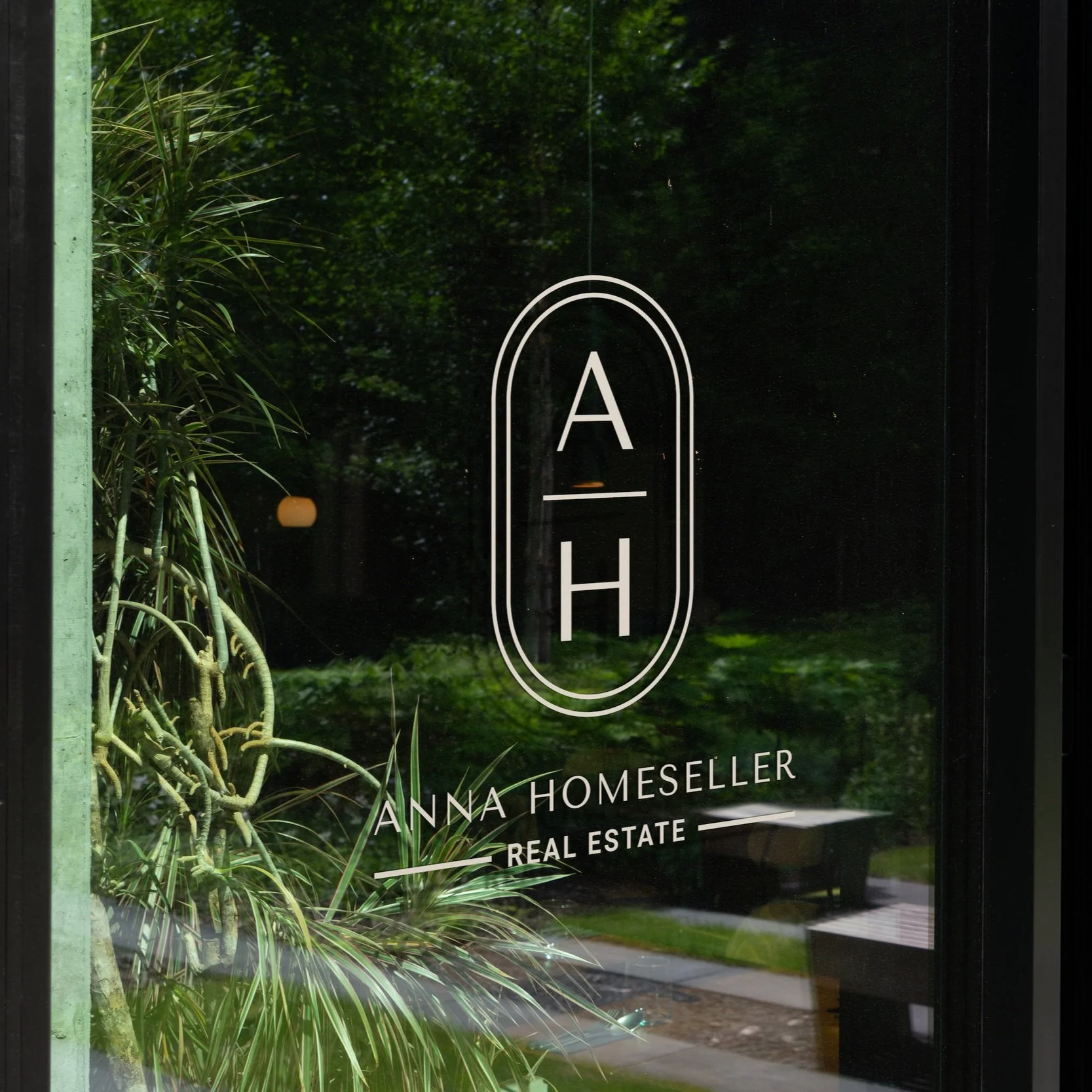 Glass window with The Anna monogram logo and plants reflecting outside.