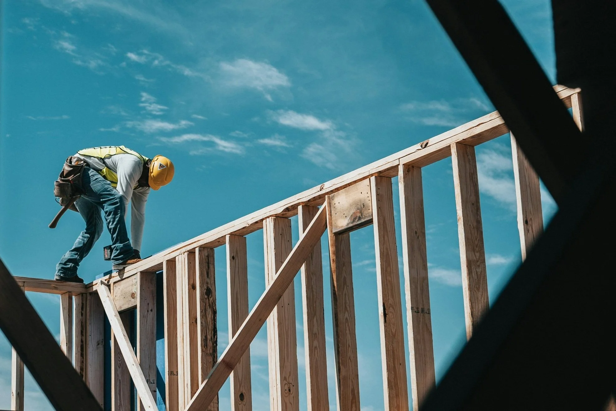 Construction worker in hard hat and safety vest standing on a wooden frame under a blue sky.