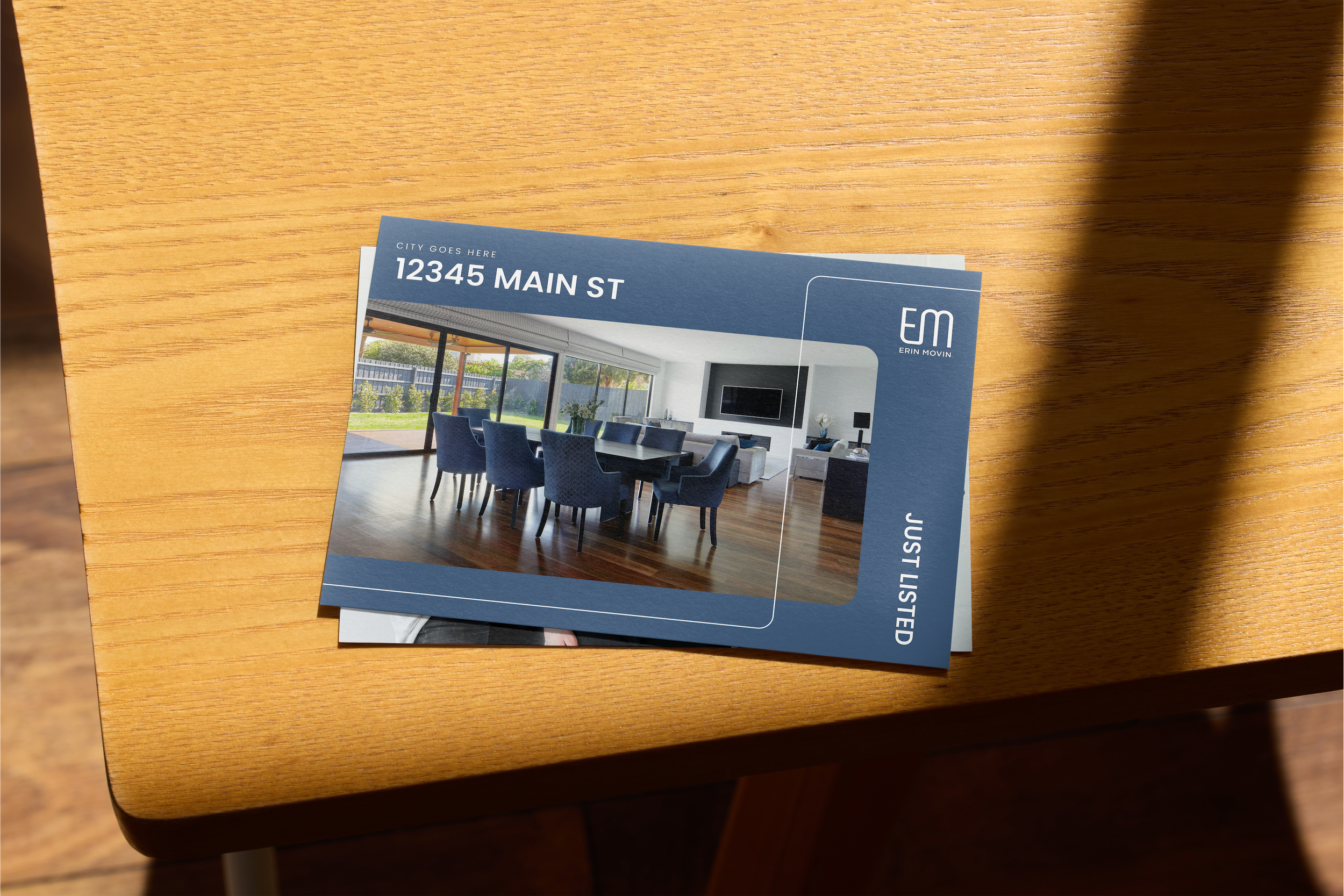 Real estate postcard on a wooden table showing a modern dining room with blue chairs, large windows, and a TV. The card reads "12345 Main St," with placeholders for city and branding information, and "Just Listed."