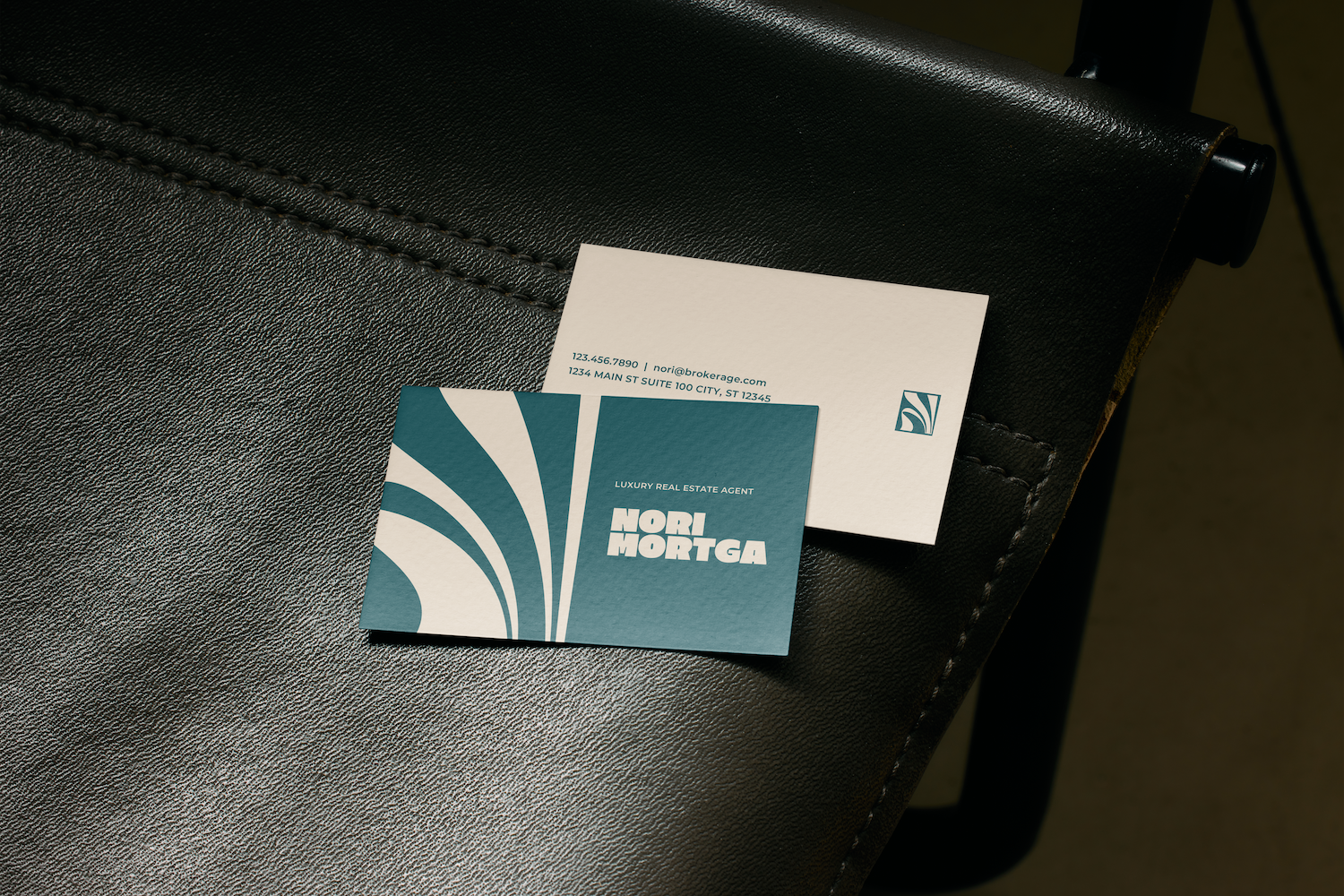 Two business cards on a black leather surface; one with teal and white abstract design and text "Luxury Real Estate Agent, Nori Mortga," the other showing contact information including a phone number, email, and address.