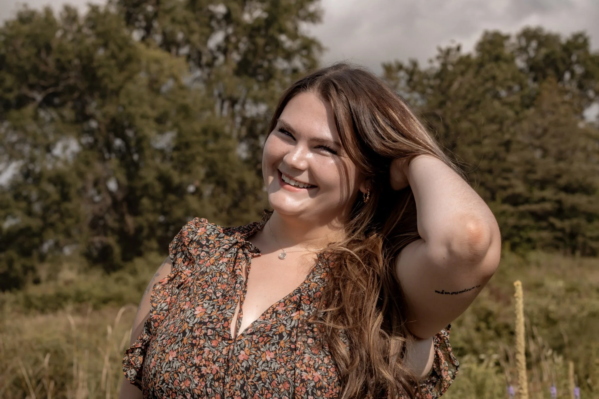 A young woman with long, wavy brown hair smiling outdoors, wearing a floral dress and a necklace, with trees in the background.